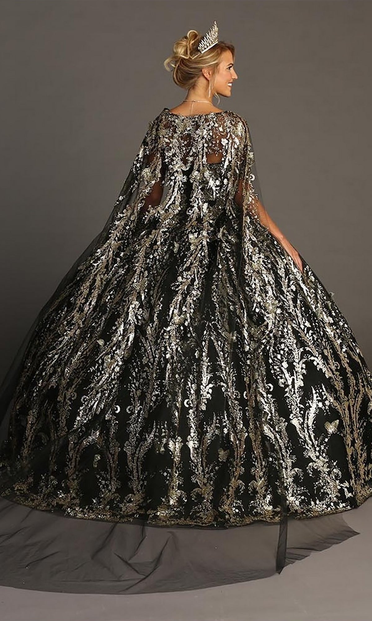 A person wearing the Quinceanera Dress CHF3101 by Chicas—featuring an off-the-shoulder neckline, a tulle ballgown skirt, metallic-silver floral embellishments, and a sheer cape—stands facing away and is adorned with a tiara.