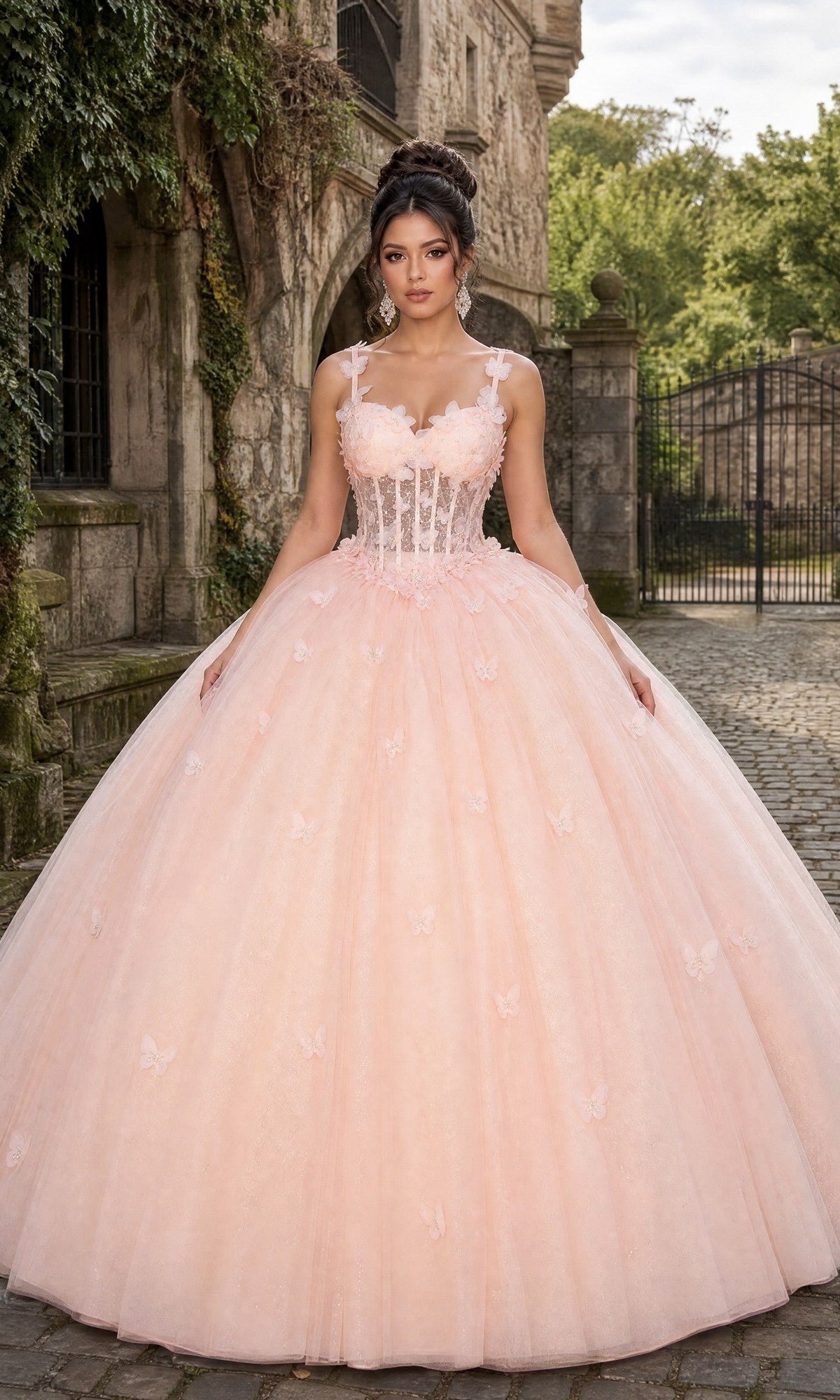 A woman stands on a cobblestone path in front of a stone building and iron gate, wearing the Quinceanera Dress CHF3107 By Chicas in blush—a ballgown with a fitted corset bodice, tulle skirt, and delicate 3D floral appliques.