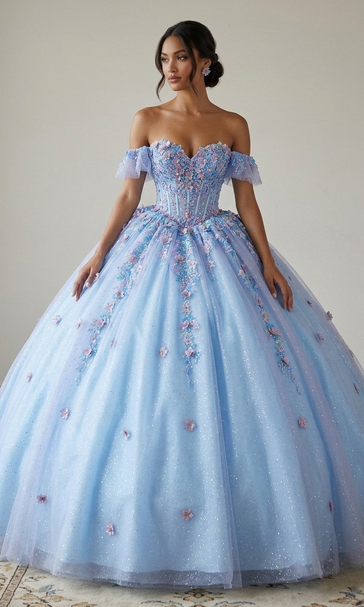 A woman stands elegantly on a patterned rug against a neutral background, wearing the Quinceanera Dress CHF3114 By Chicas in blue, featuring an off-the-shoulder design, fitted bodice with 3D floral appliques, and a full sparkly skirt.