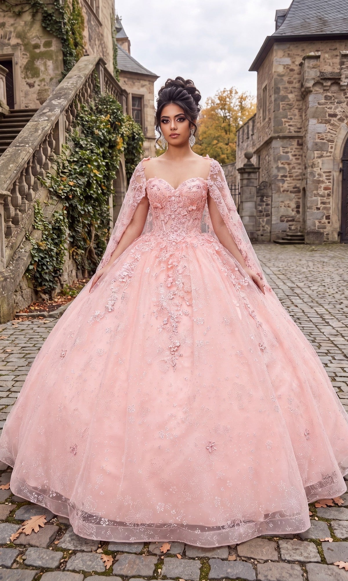 Wearing the Quinceanera Dress CHF3118 By Chicas in baby pink, a woman stands on a cobblestone path between stone buildings, surrounded by greenery climbing the walls and glowing autumn trees. The gown’s voluminous ballgown skirt and delicate 3D floral appliques add an enchanting touch to the scene.