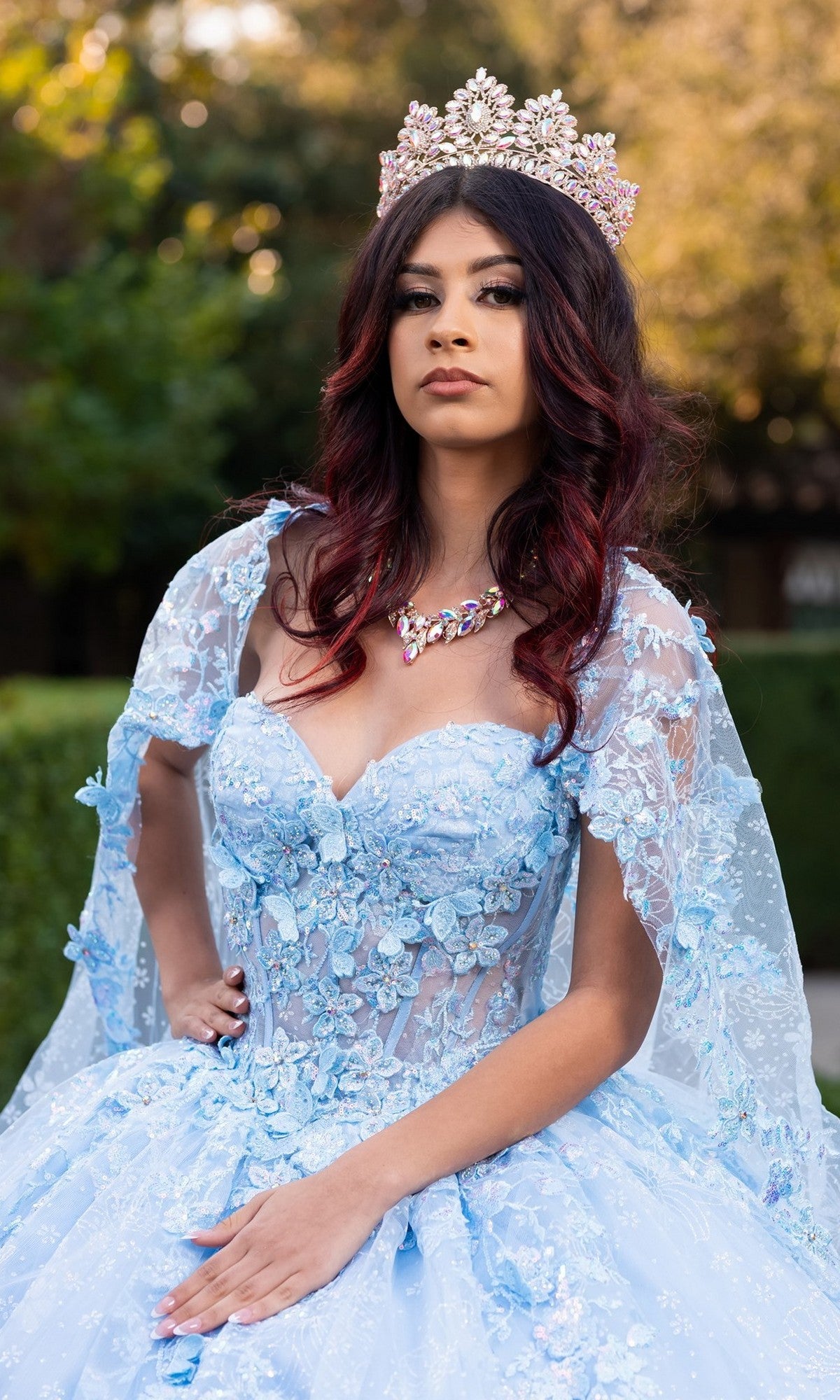 Wearing the Quinceanera Dress CHF3118 By Chicas—a blue floral ballgown skirt adorned with delicate 3D floral appliques—a woman stands outdoors amidst lush greenery, accessorized with a jeweled crown and necklace.