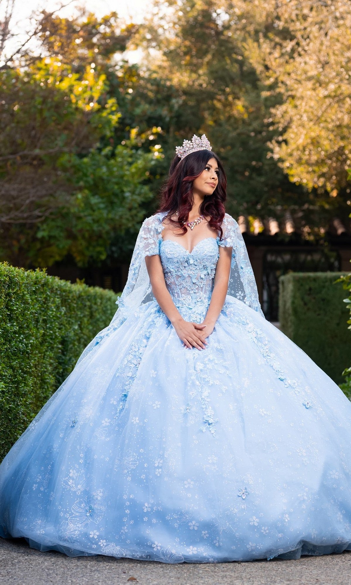 A young woman stands outdoors in the Quinceanera Dress CHF3118 By Chicas, featuring a pastel hue with 3D floral appliques and a voluminous ballgown skirt. With her hands clasped, she gazes to the side as her shimmering crown rests atop her head.