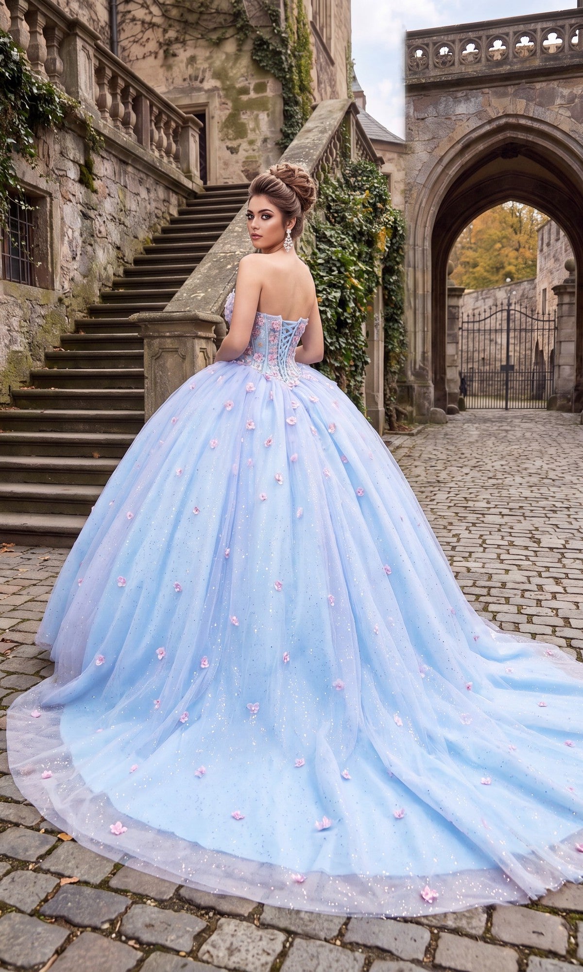 A woman wearing the Quinceanera Dress CHF3119 By Chicas, a strapless ballgown adorned with pink 3D floral appliques, stands on a cobblestone path near stone stairs and an old building.