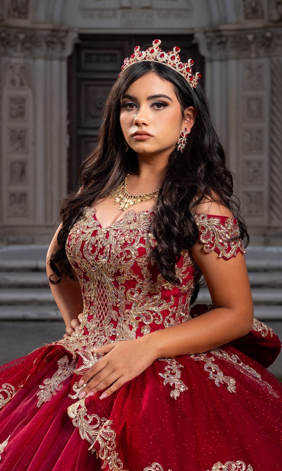 A young woman stands gracefully in front of ornate stone steps and a decorative doorway, wearing the Quinceanera Dress CHF3120 By Chicas—a stunning red and gold off-the-shoulder gown adorned with delicate gold embroidery and glitter tulle details—her look completed with a matching crown and elegant jewelry.