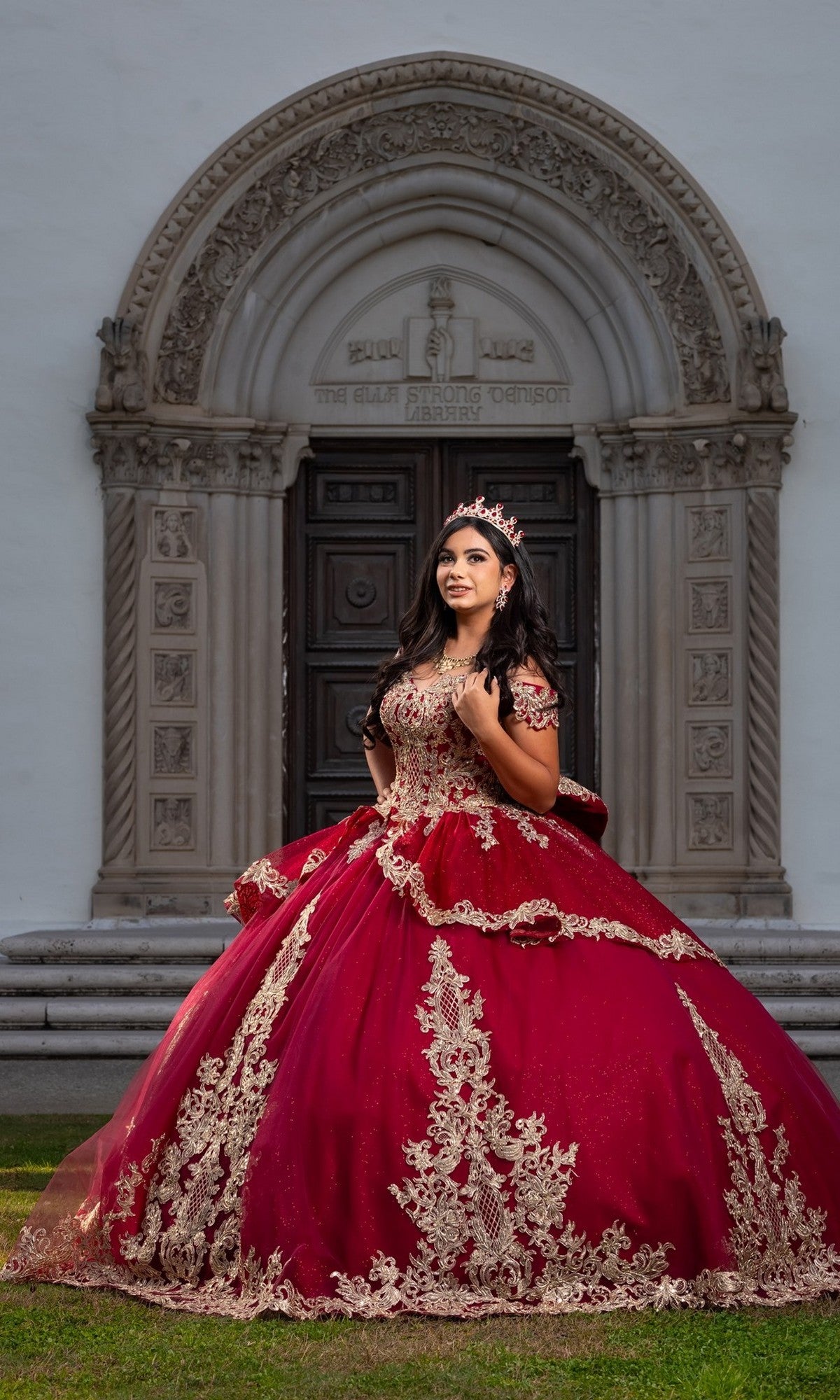 A young woman wearing the Quinceanera Dress CHF3120 By Chicas, an off-the-shoulder ball gown adorned with gold embroidery, stands on grass before an ornate arched doorway, her red and gold dress shimmering beneath a delicate tiara.