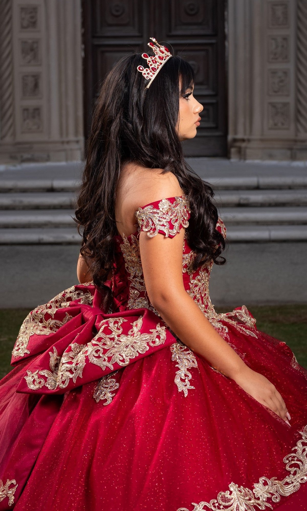 A young woman sits in profile outdoors, gracefully posing before a large building with carved doors, her Quinceanera Dress CHF3120 By Chicas shimmering with ornate gold embroidery and delicate red and gold details, complemented by a sparkling tiara.