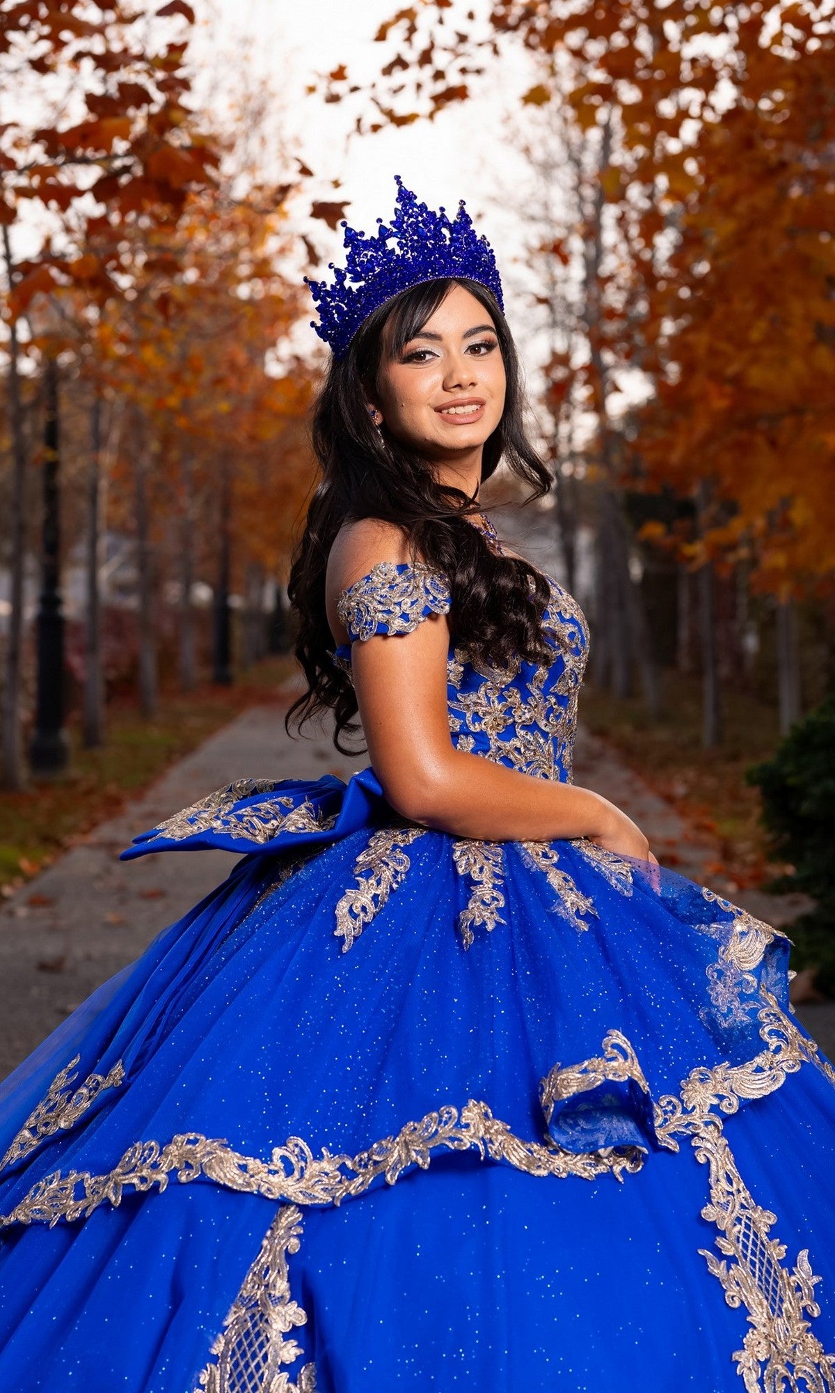 A young woman in the Quinceanera Dress CHF3120 By Chicas and a matching crown stands on a tree-lined path with autumn leaves in the background.
