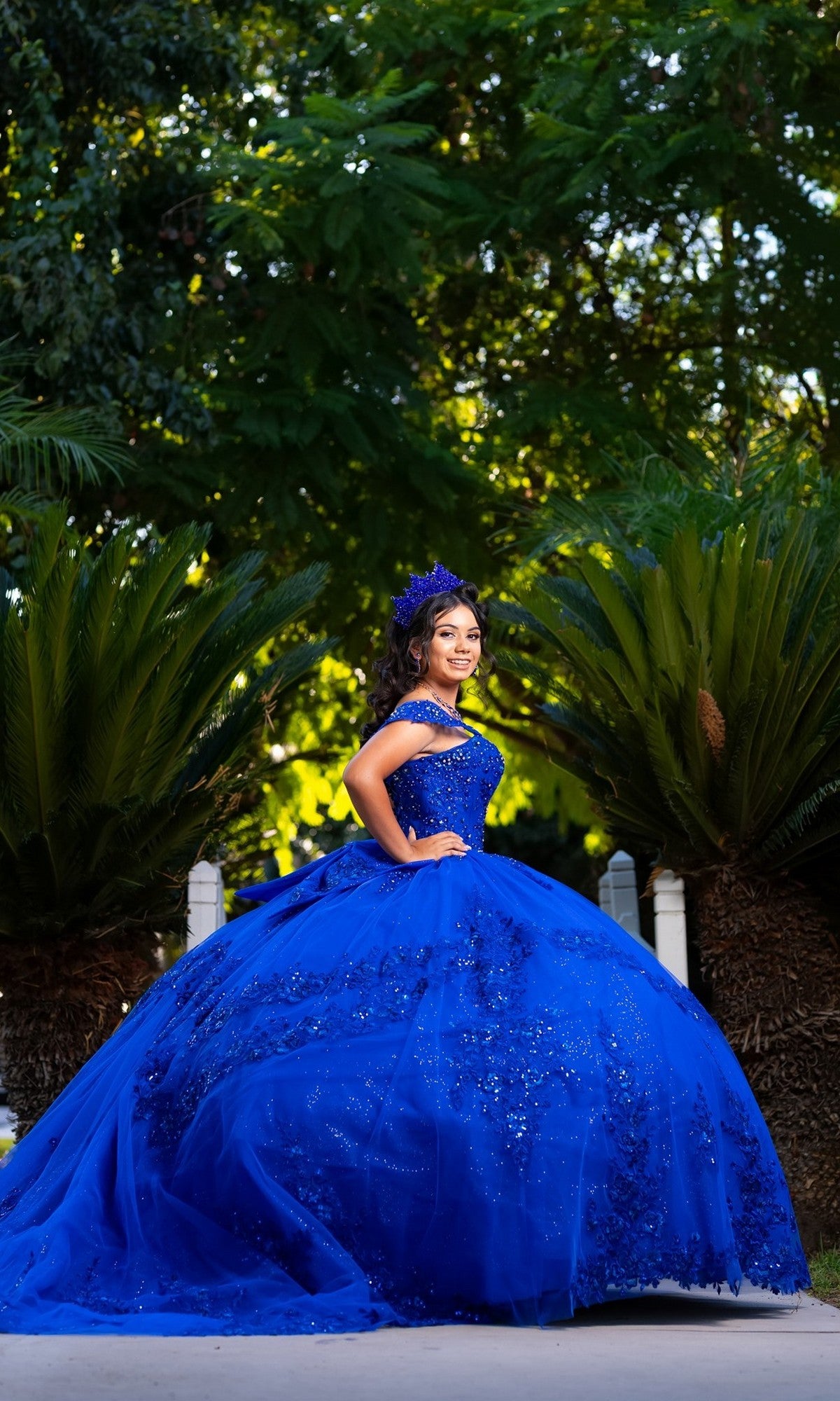 A young woman in the Quinceanera Dress CHF3122 by Chicas, a royal blue off-the-shoulder gown, poses outdoors against vibrant green foliage, gazing confidently at the camera.