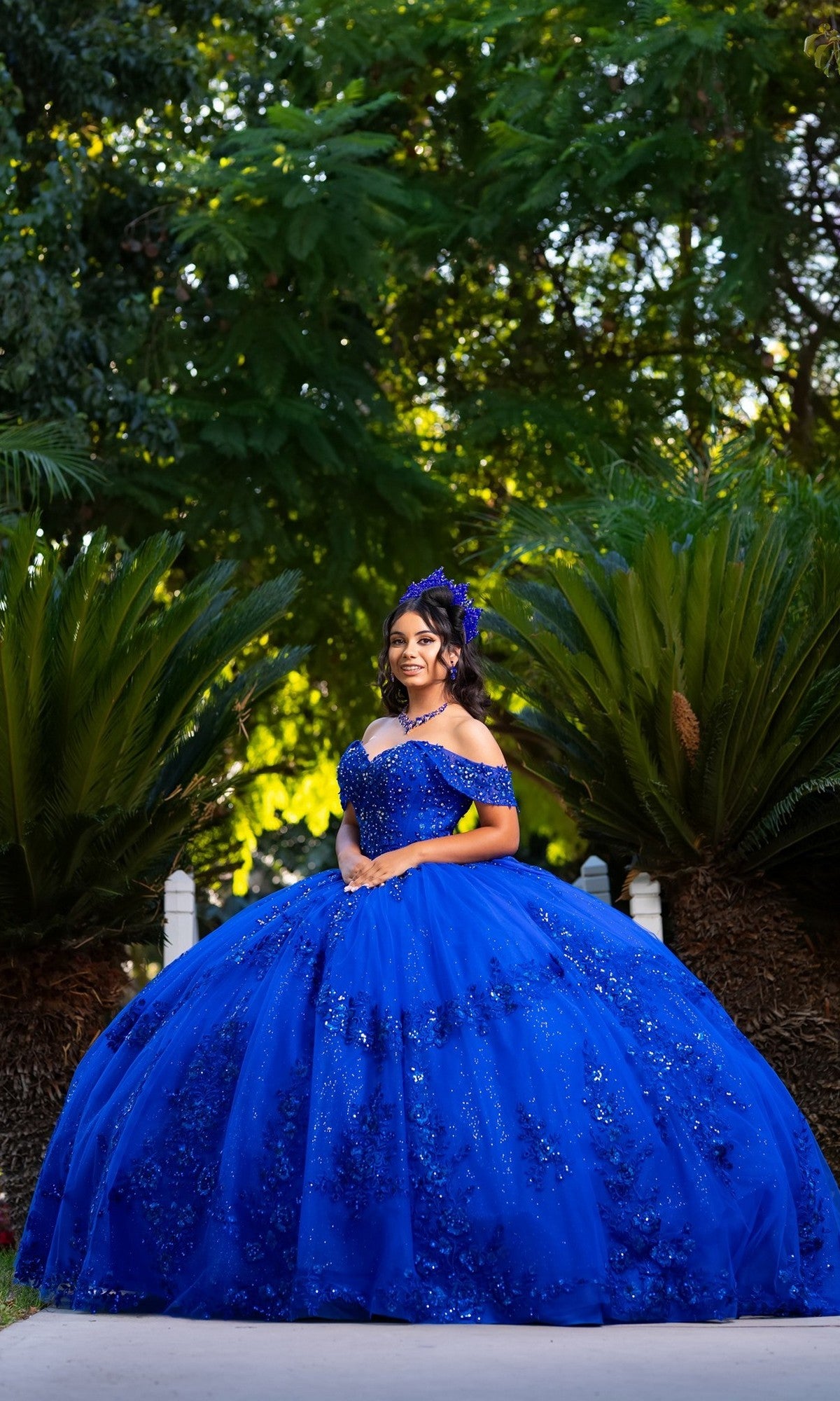 A young woman in the Quinceanera Dress CHF3122 By Chicas, featuring an off-the-shoulder design and elaborate royal blue detailing, stands outdoors among lush green foliage and palm-like plants, looking at the camera with elegance.