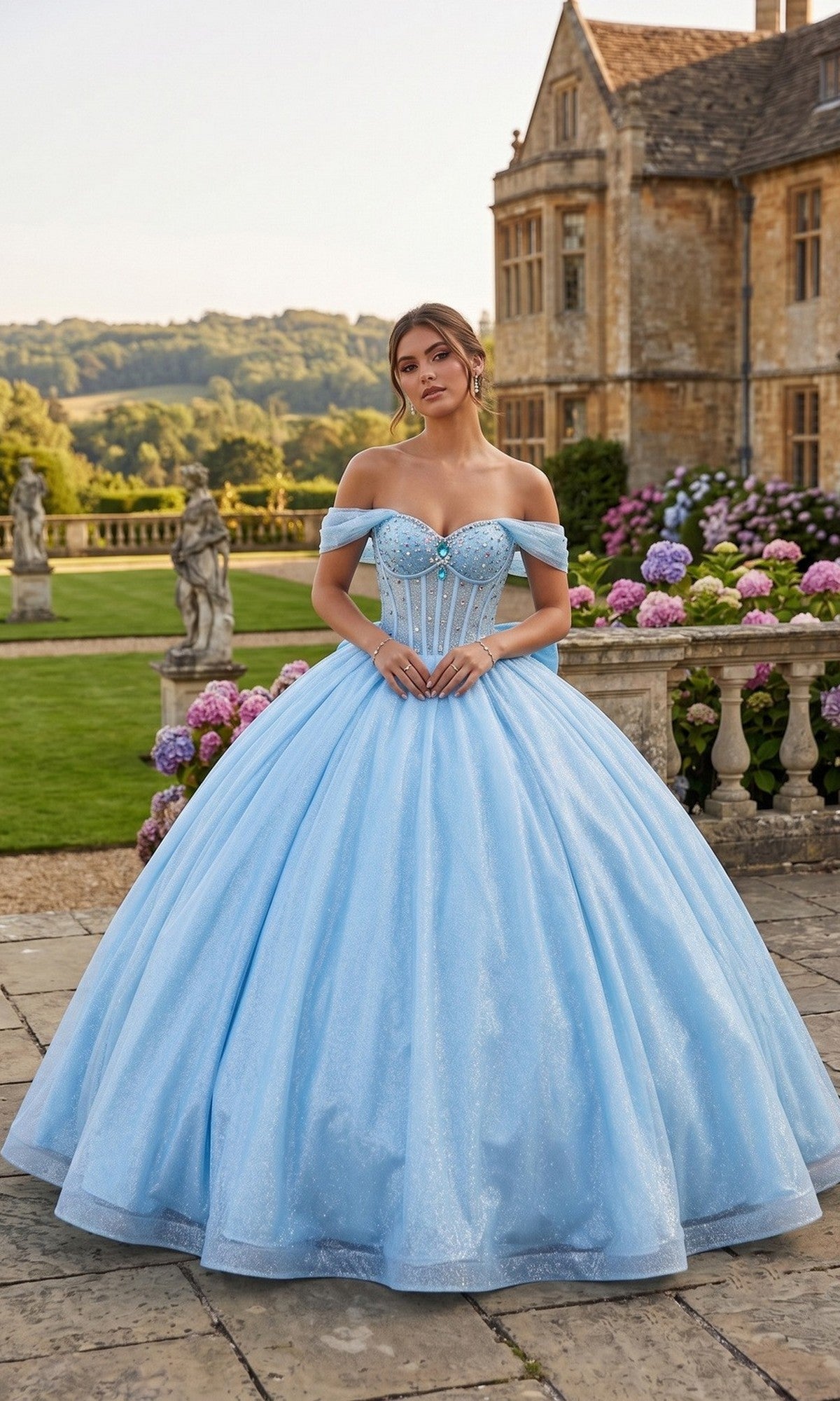A woman in the Quinceanera Dress CHF3125 by Chicas, a baby blue off-the-shoulder glitter-tulle ballgown with a delicate corset bodice, stands on a stone patio in front of a historic mansion and colorful hydrangea bushes.