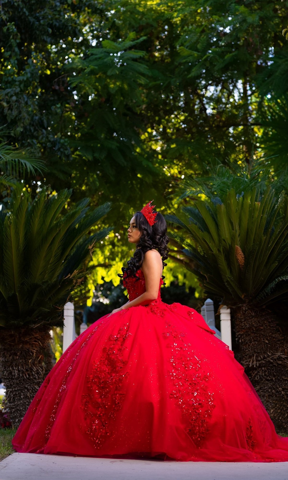A young woman stands outdoors between two large palm plants, surrounded by lush greenery, wearing the Quinceanera Dress CHF3129 By Chicas—a bright red strapless ballgown that shimmers with layers of glitter tulle. Her outfit is complemented by a matching headpiece, adding a magical touch to the vibrant scene.