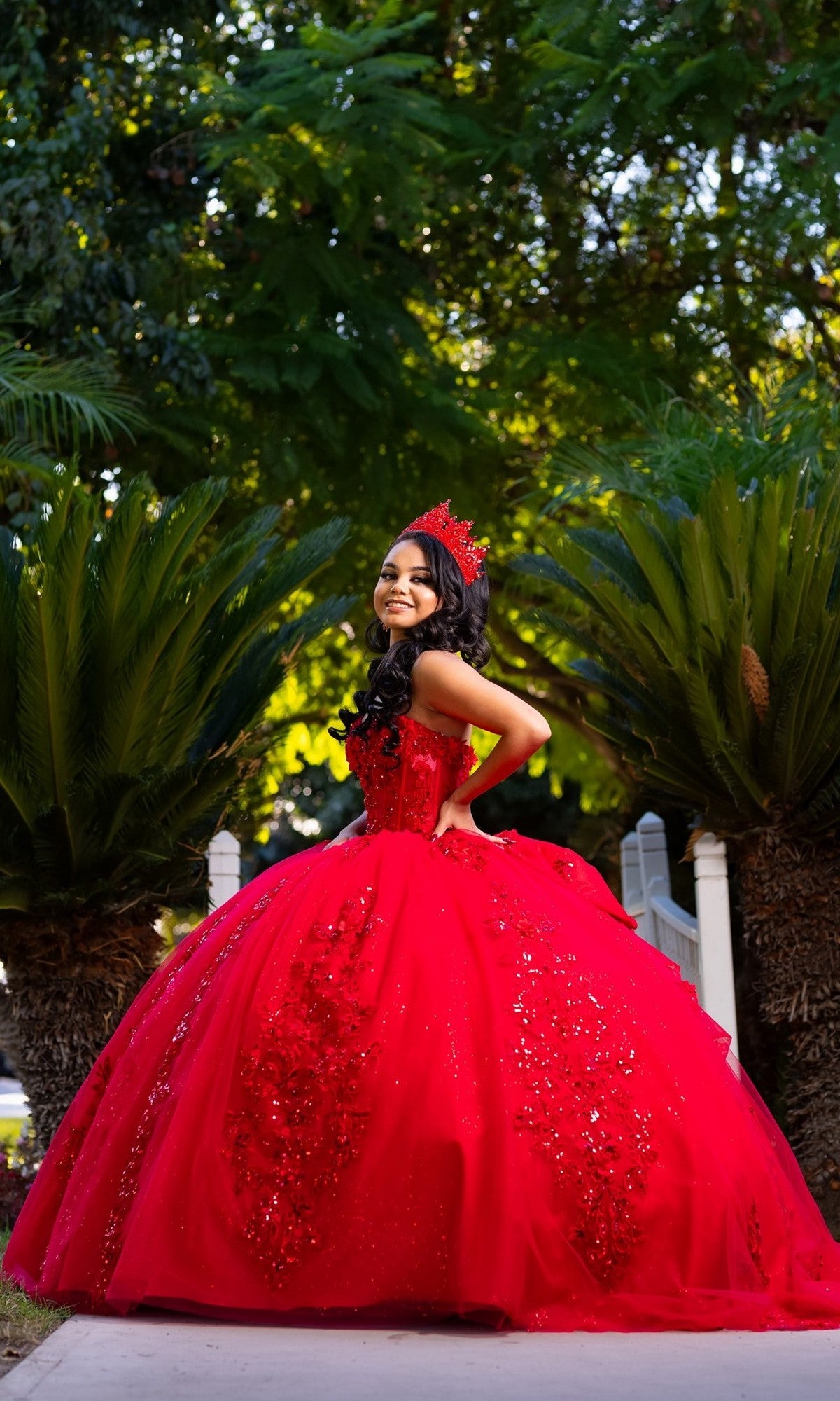 A young woman in the Quinceanera Dress CHF3129 By Chicas—a bright red, strapless ballgown adorned with a matching crown—stands outdoors, smiling amid lush greenery. The gown’s layers of glitter tulle shimmer beautifully in the natural light.