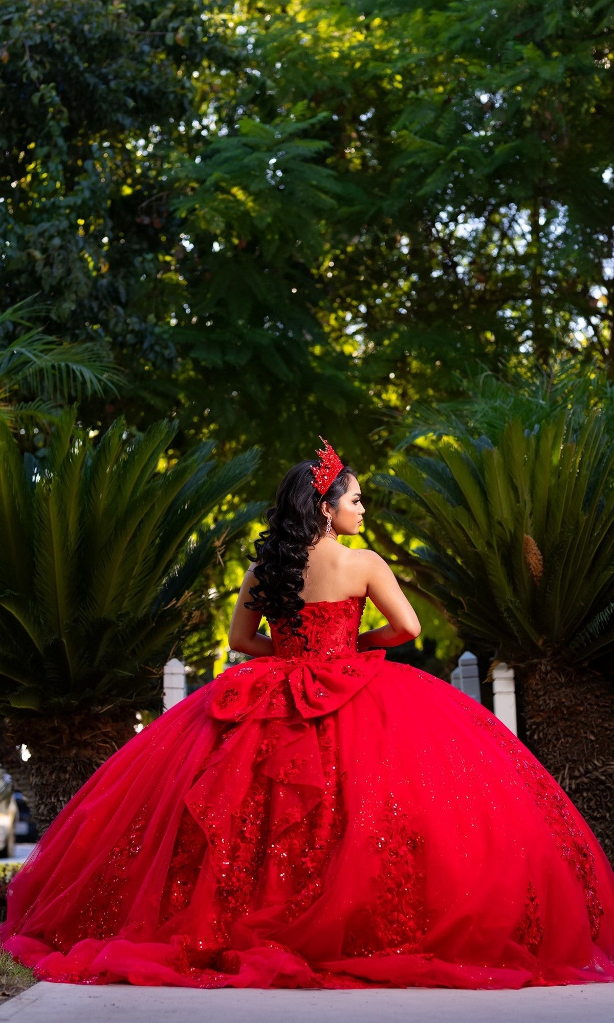 A young woman wearing the Quinceanera Dress CHF3129 By Chicas, featuring sparkling red glitter tulle and a matching crown, stands outdoors facing away, surrounded by green foliage.