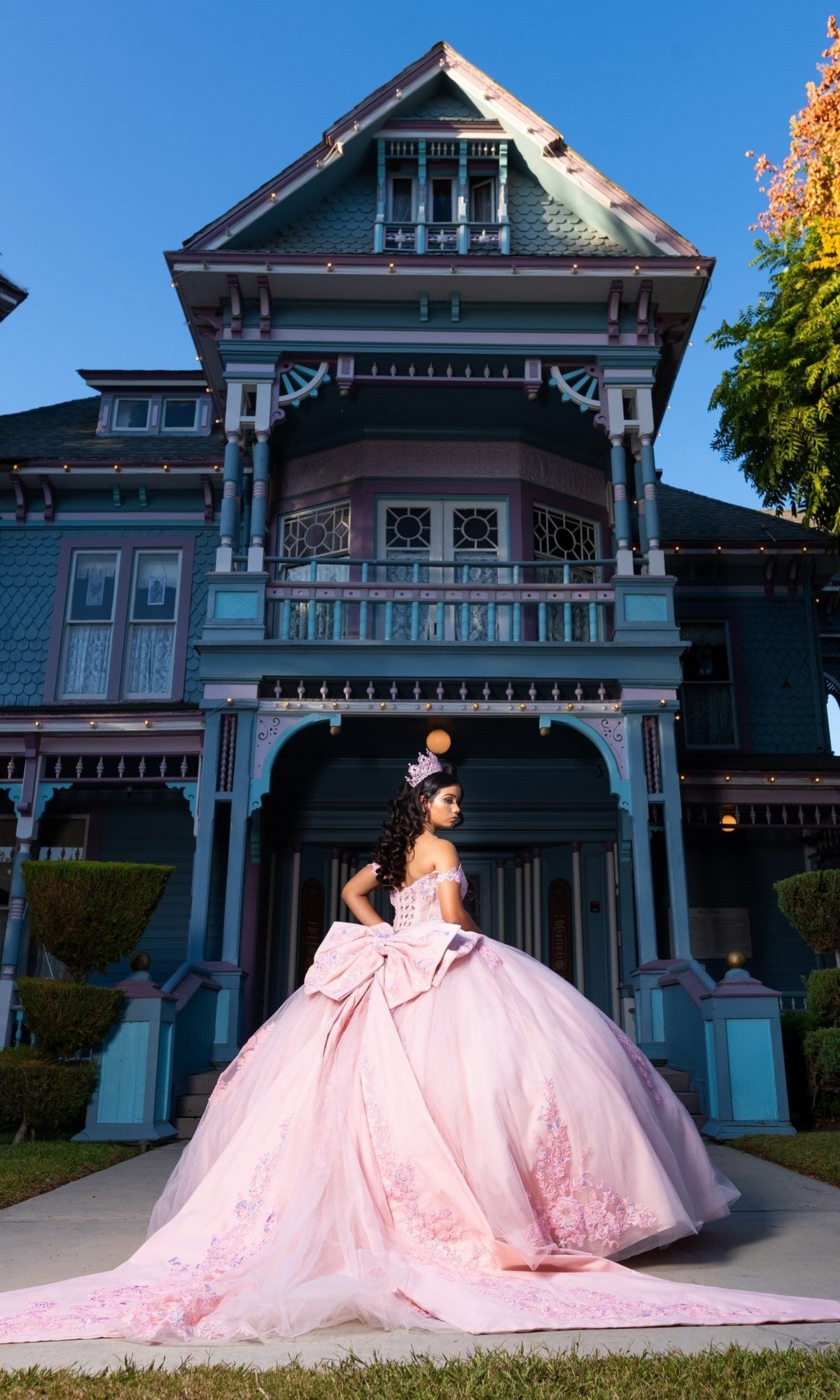 A young woman wearing the Quinceanera Dress CHF3130 by Chicas, featuring an off-the-shoulder corset-style bodice and a tulle ballgown skirt, stands in front of a grand Victorian-style house on a sunny day, her shimmering pink gown and delicate tiara catching the light.