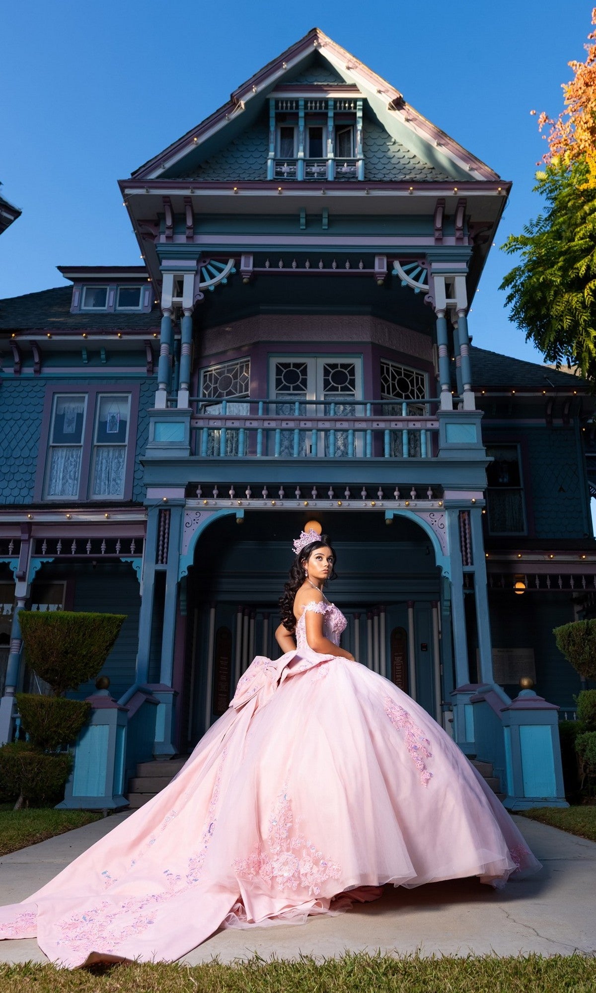 A young woman wearing the Quinceanera Dress CHF3130 by Chicas, a pink off-the-shoulder gown with a corset-style bodice and a sweeping tulle ballgown skirt, stands in front of a large, ornate Victorian-style house on a sunny day.
