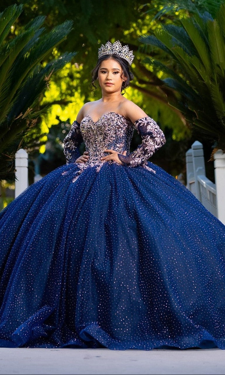 A young woman in the Quinceanera Dress CHF3131 By Chicas in navy, adorned with a jeweled crown, stands on a bridge surrounded by lush green foliage.