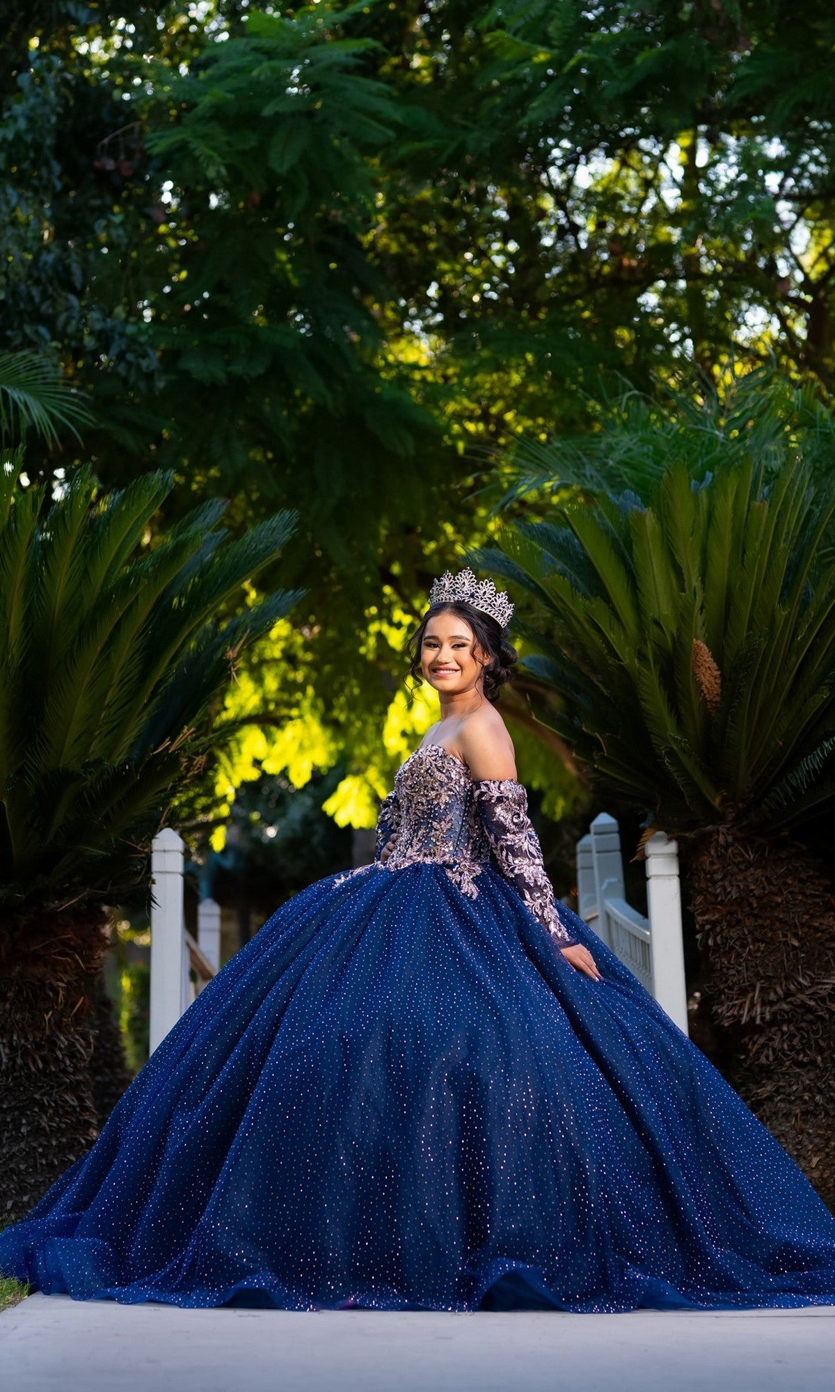 A young woman in the Quinceanera Dress CHF3131 By Chicas and a crown stands outdoors, smiling, with greenery and trees in the background.