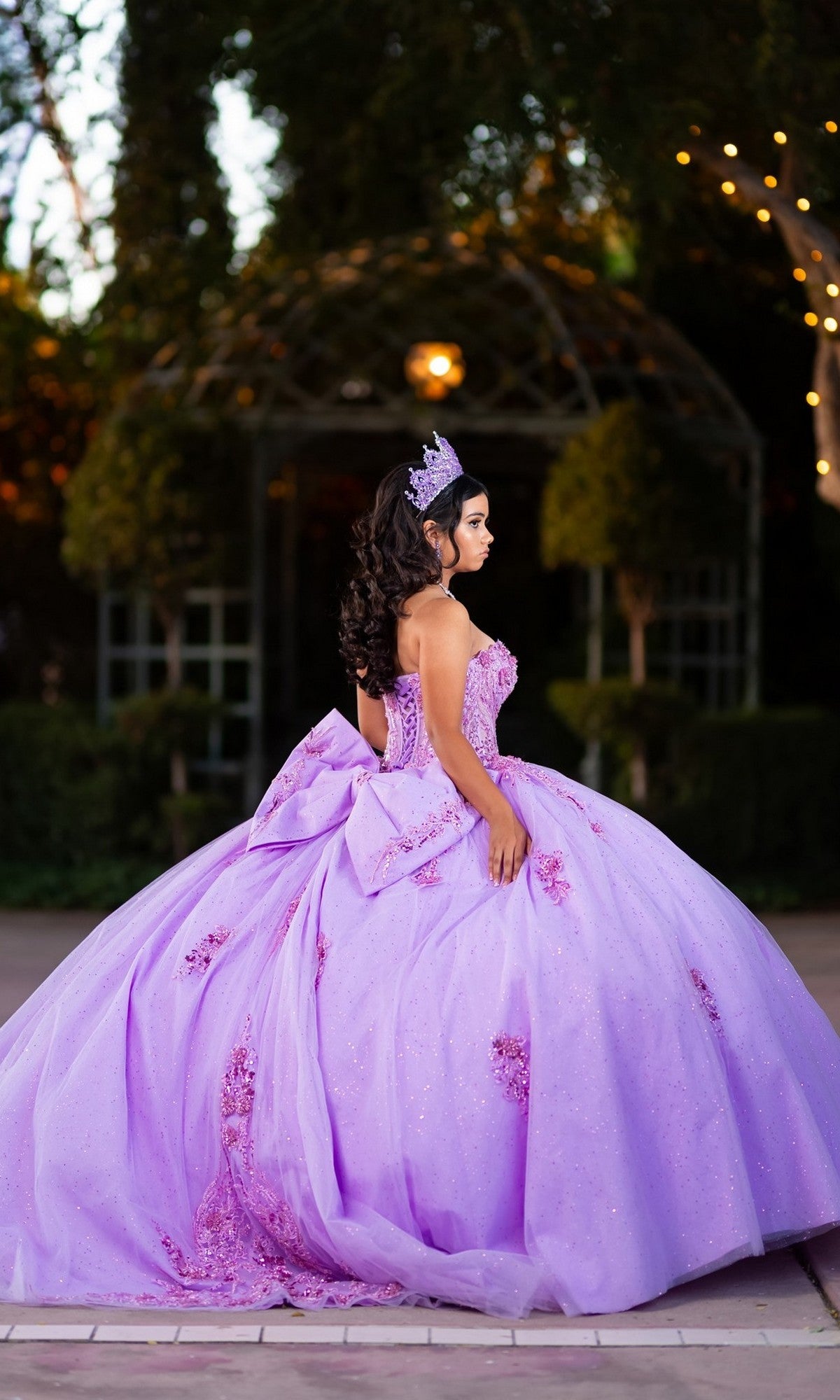 A young woman stands outdoors, facing sideways, wearing the Quinceanera Dress CHF3133 By Chicas—a lavender glitter tulle ballgown with a corset-style bodice and tiara—framed by trees and a gazebo in the background.