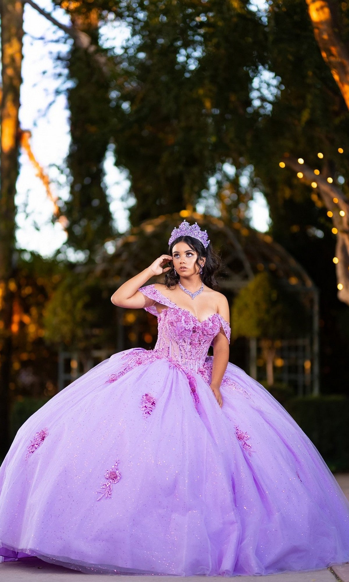 A young woman wearing the Quinceanera Dress CHF3133 By Chicas—a sparkling tulle ballgown with a delicate corset bodice—poses outdoors, surrounded by trees and glowing string lights, her look completed with a shimmering tiara.