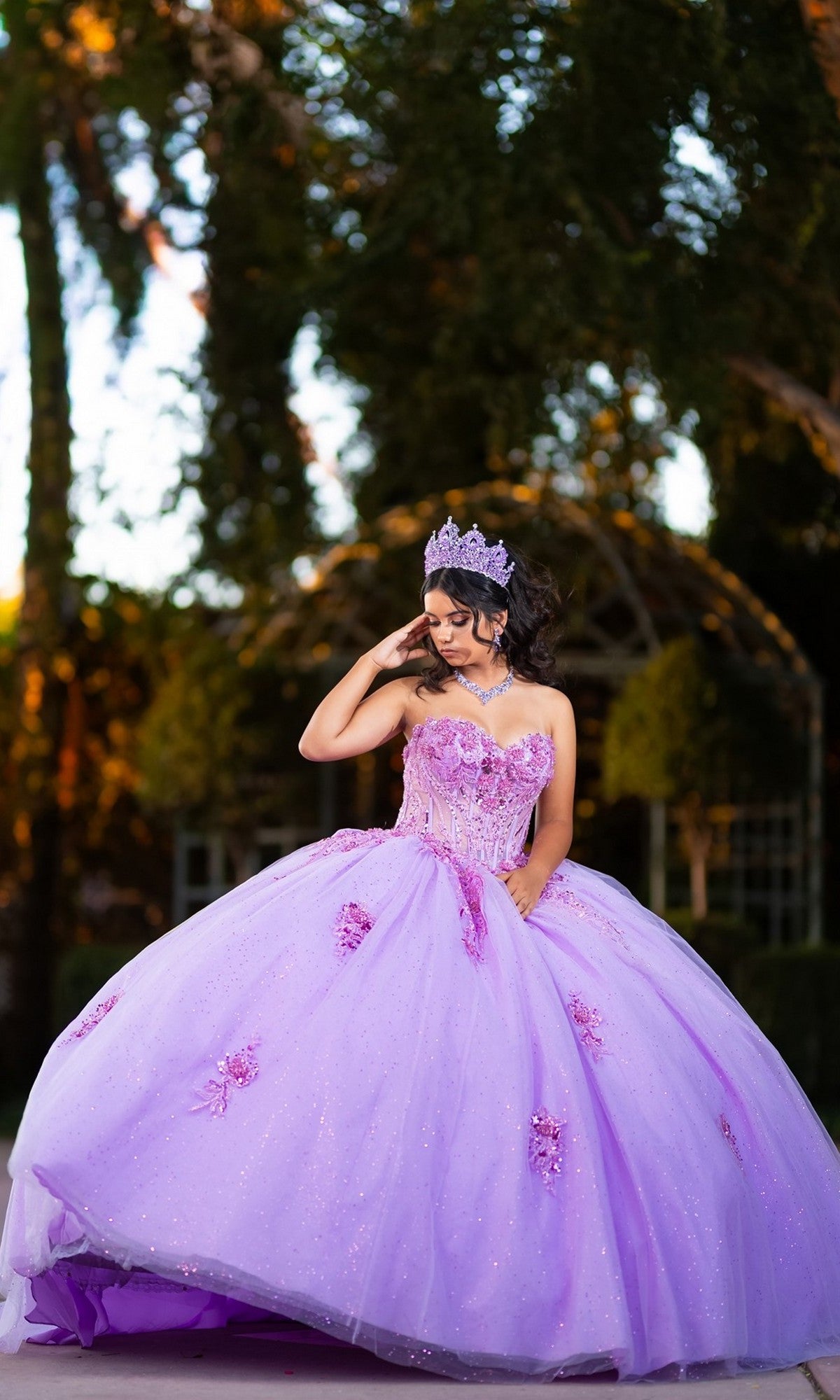 A young woman stands outdoors, looking down with one hand gently touching her face, wearing the Quinceanera Dress CHF3133 by Chicas—a sparkling lavender glitter tulle ballgown paired with a tiara.