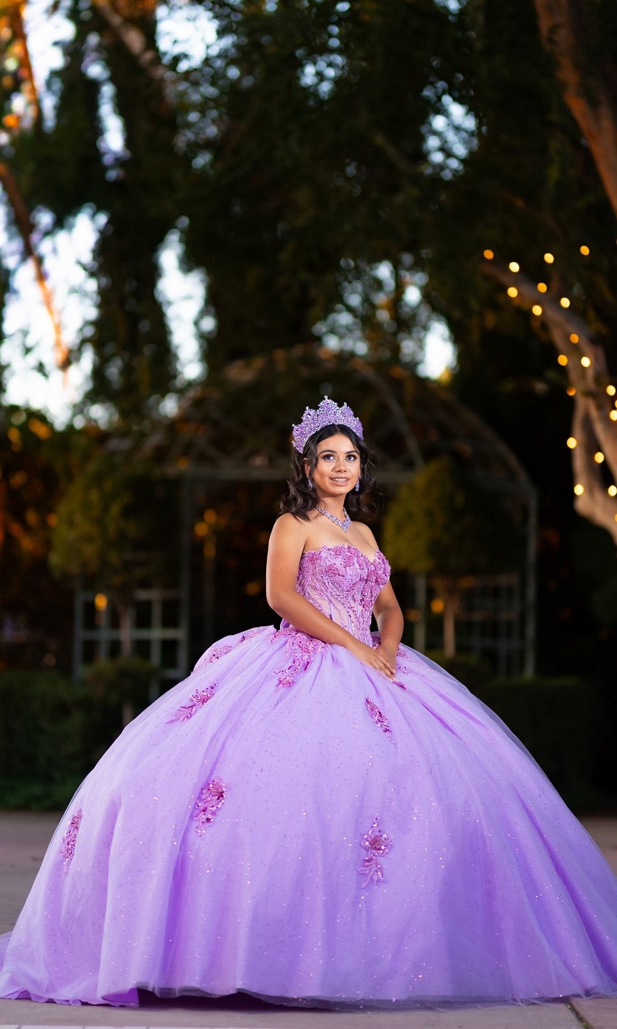 A young woman stands outdoors surrounded by trees and soft string lights, wearing the Quinceanera Dress CHF3133 By Chicas—a lavender glitter tulle ballgown with a corset bodice—accented by a jeweled crown.
