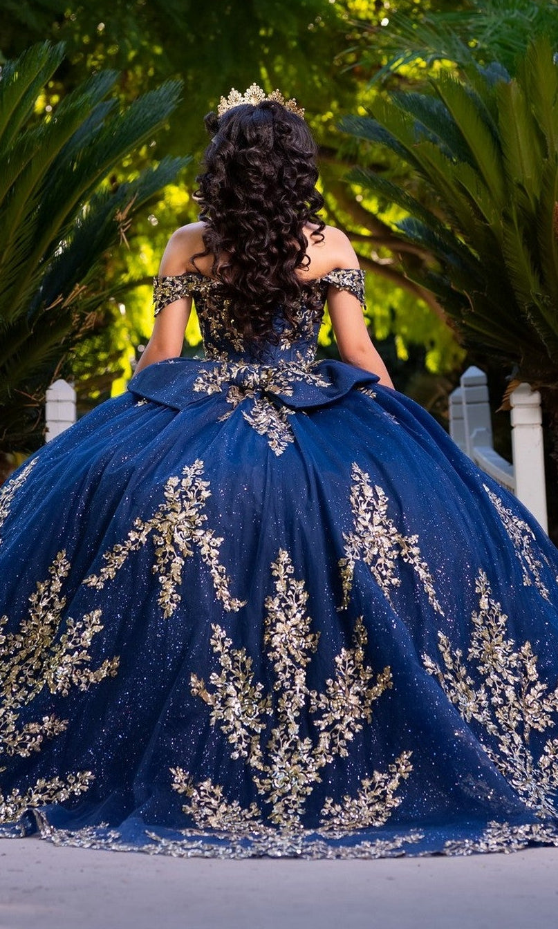 A person with long, curled hair is seen standing outdoors among green plants, wearing the Quinceanera Dress CHF3134 By Chicas—an off-the-shoulder gown adorned with gold embroidery and paired with a crown.