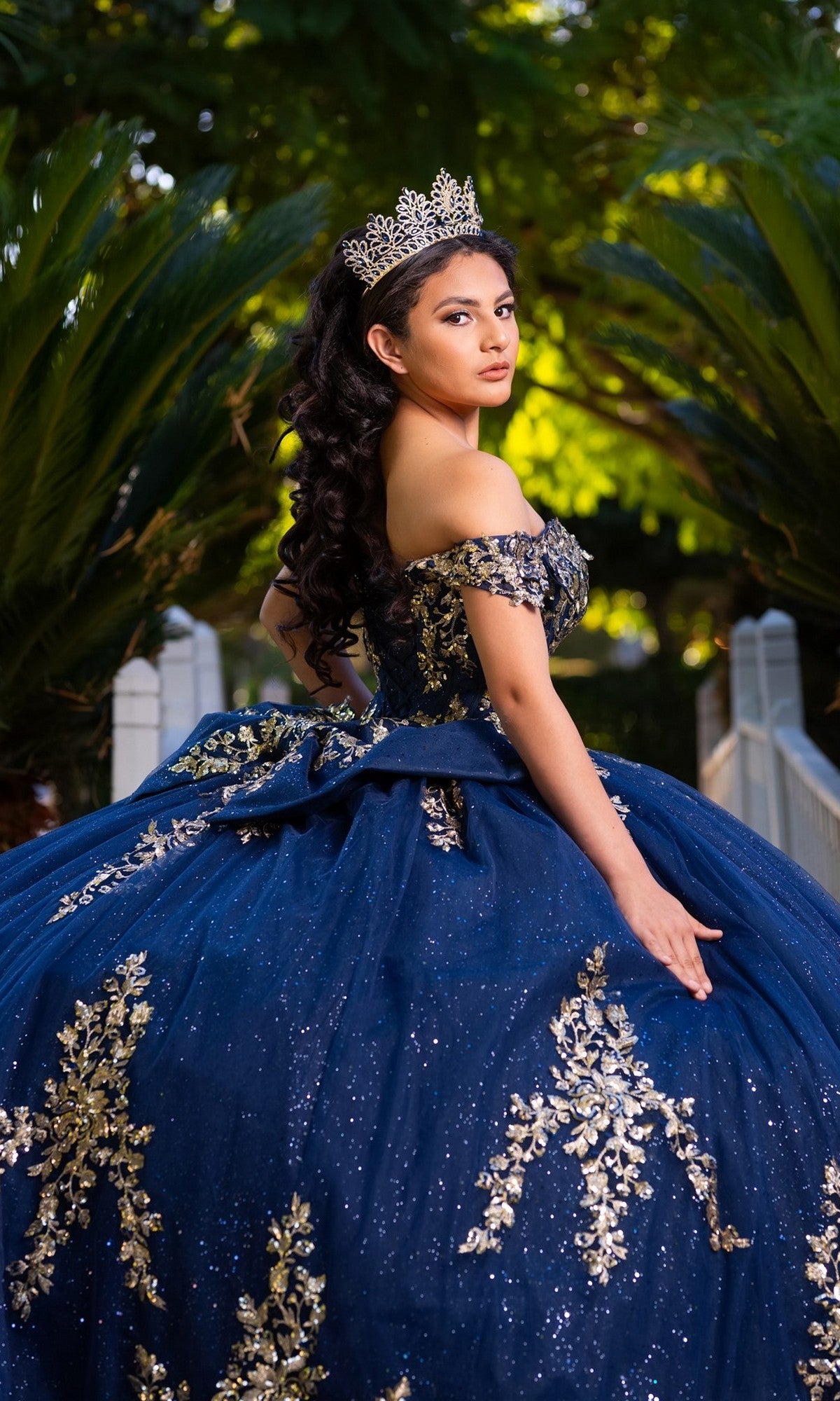 A young woman stands outdoors framed by greenery, looking over her shoulder in the Quinceanera Dress CHF3134 By Chicas—a stunning blue and gold gown featuring off-the-shoulder sleeves and delicate gold embroidery.