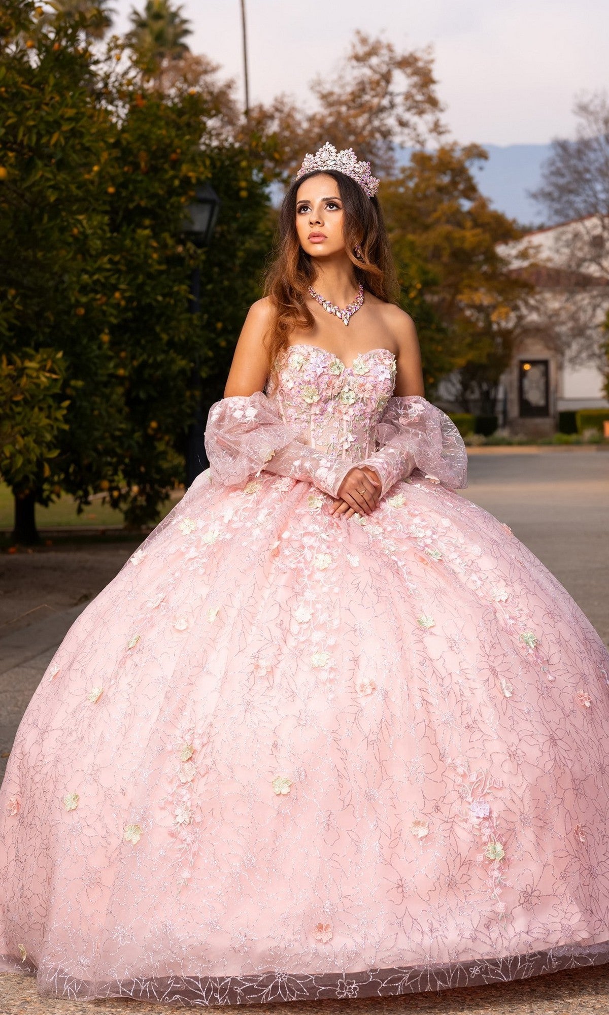A young woman wearing the Quinceanera Dress CHF3140 By Chicas, featuring 3D floral appliques and a tiara, stands outdoors in her pastel ballgown skirt, posing for a formal portrait with trees and buildings in the background.
