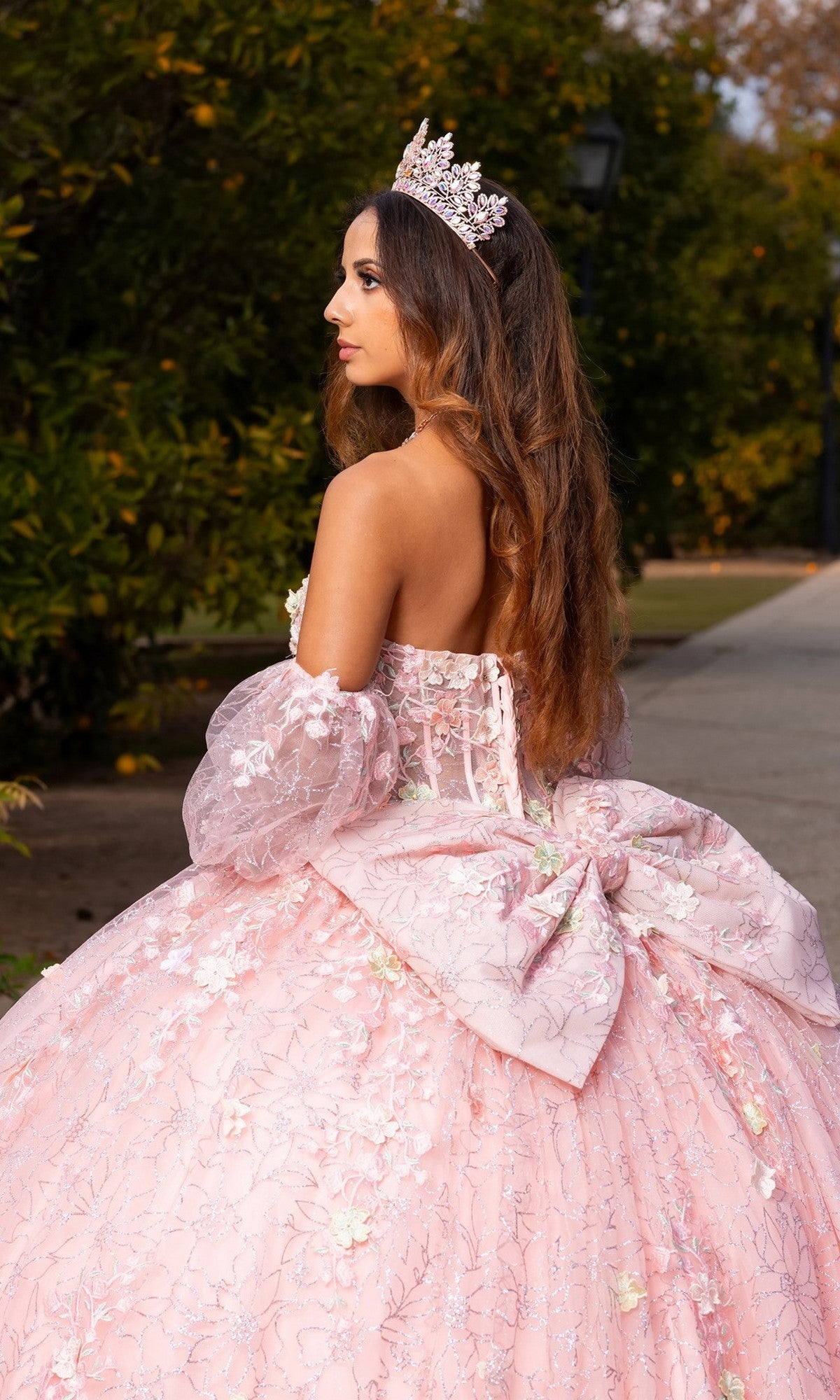 A young woman stands outdoors in the Quinceanera Dress CHF3140 By Chicas—a pastel ballgown adorned with delicate 3D floral appliques and paired with a jeweled crown—looking elegantly to the side.