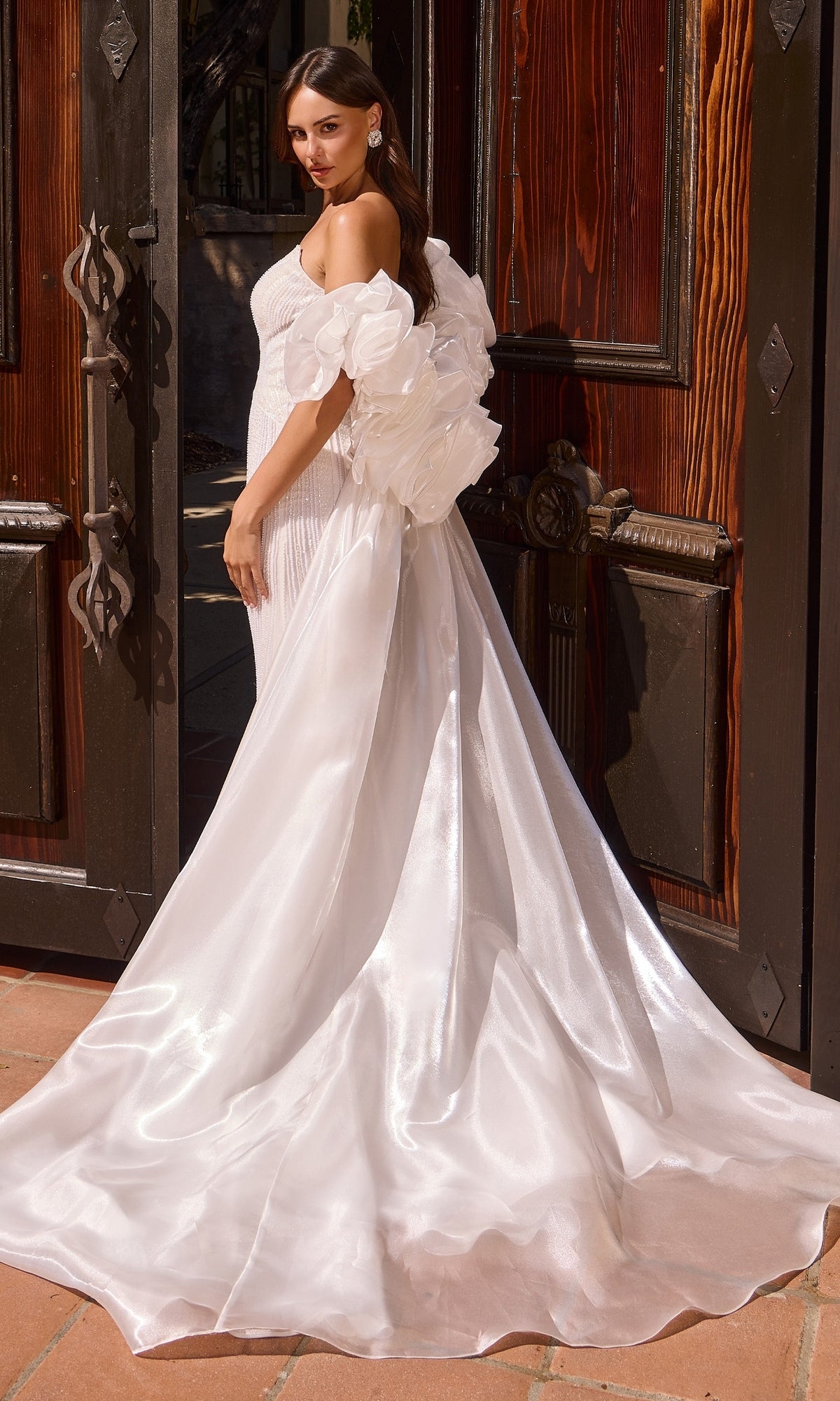 A woman stands in front of wooden doors wearing the Ladivine CJ933W long prom dress, featuring a white off-shoulder design with a mermaid skirt, long train, sweetheart neckline, and large floral accents.