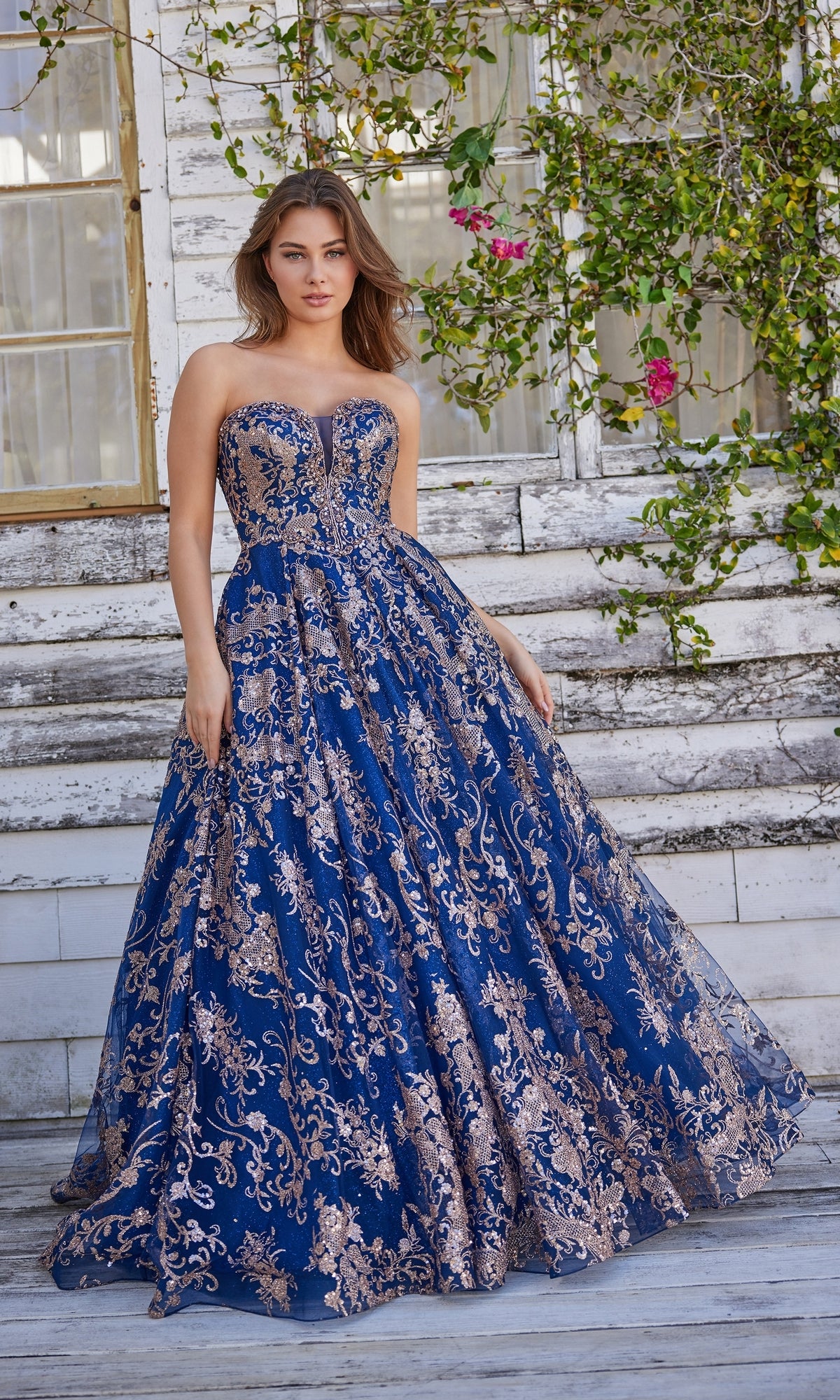 A woman in a Colette Strapless Long Prom Ball Gown CL5101, floor-length blue with gold embroidery, stands on wooden steps before a weathered building decorated with climbing plants and flowers.
