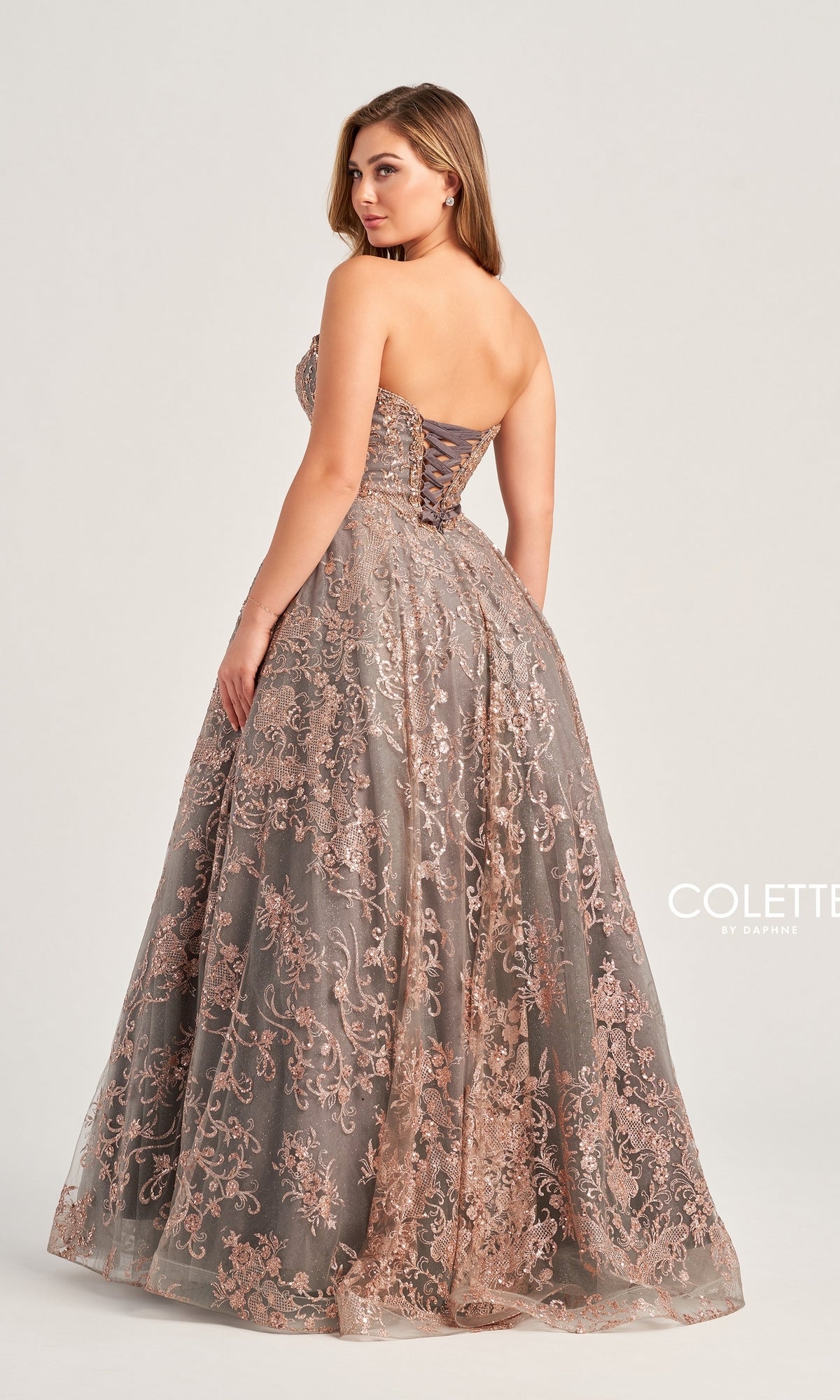 A woman stands with her back to the camera, wearing the elegant Colette Strapless Long Prom Ball Gown CL5101, which features intricate pink lace details and a chic lace-up back.