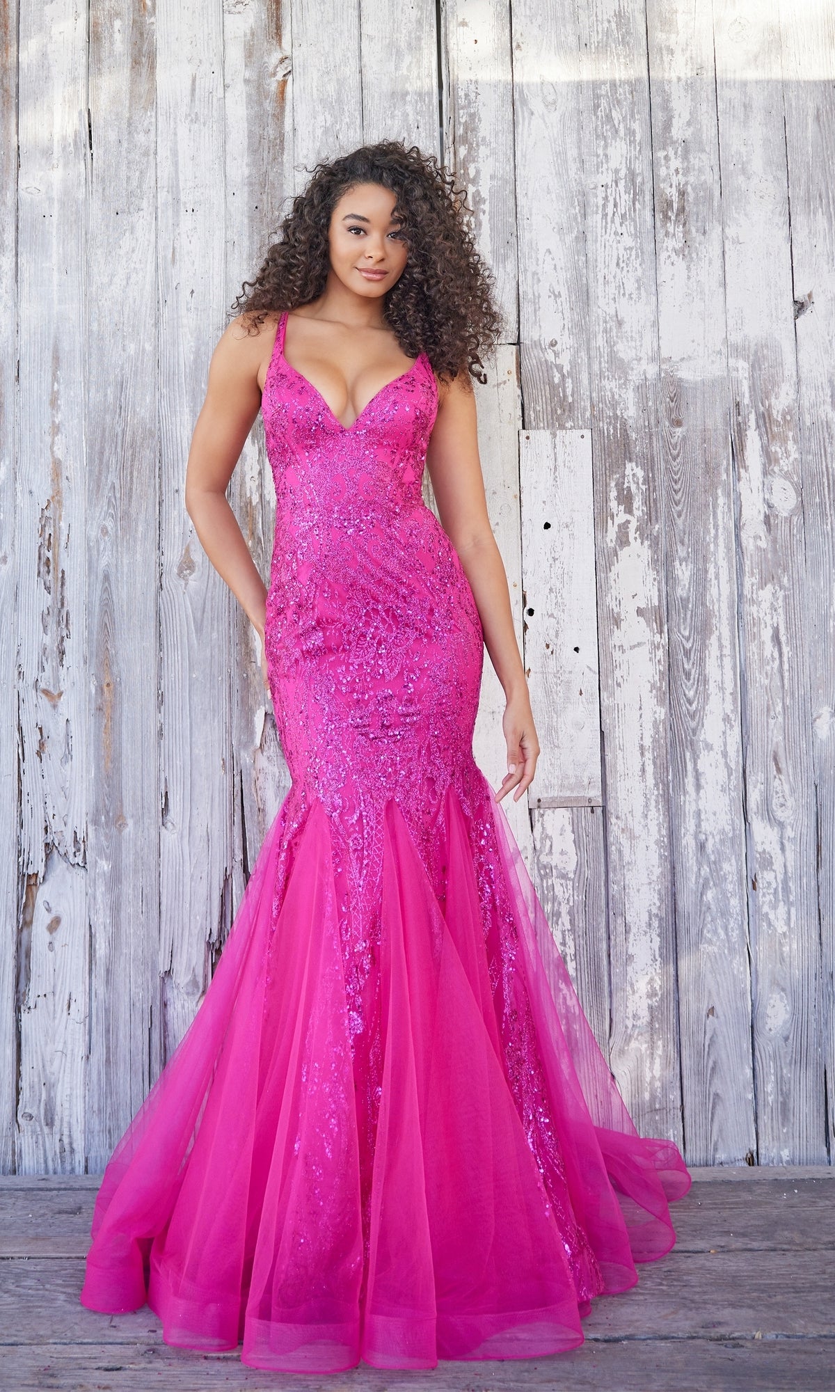 A woman wearing a bright pink, sequined Colette Long Glitter Mermaid Prom Dress CL5109 with tulle details stands in front of a weathered wooden wall.