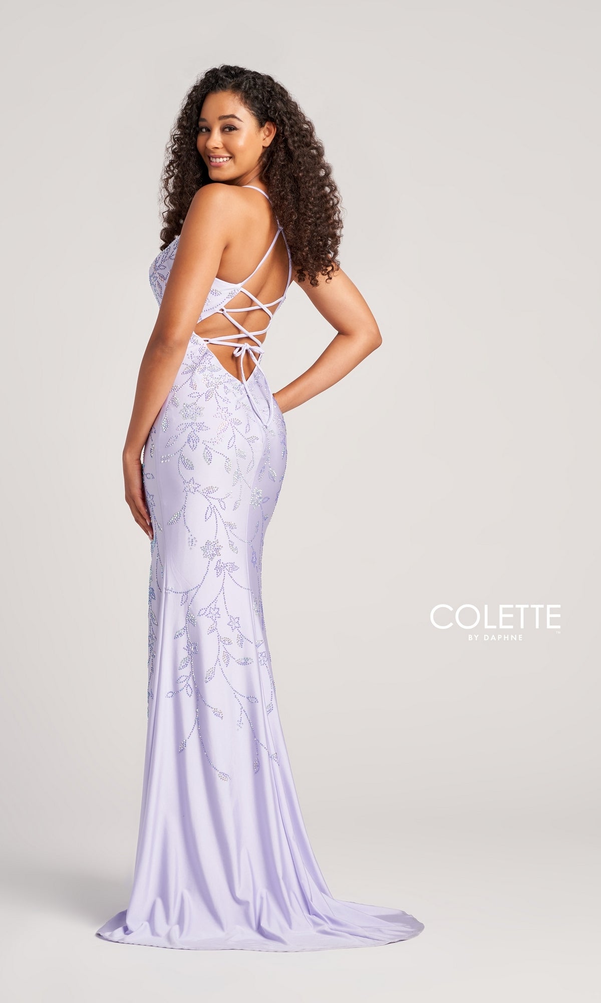 A woman in the Glitter-Print Long Colette Prom Dress CL5110 stands sideways and smiles, showcasing its light purple hue, beaded floral print, lace-up back, and floor-length design against a plain light gray background.