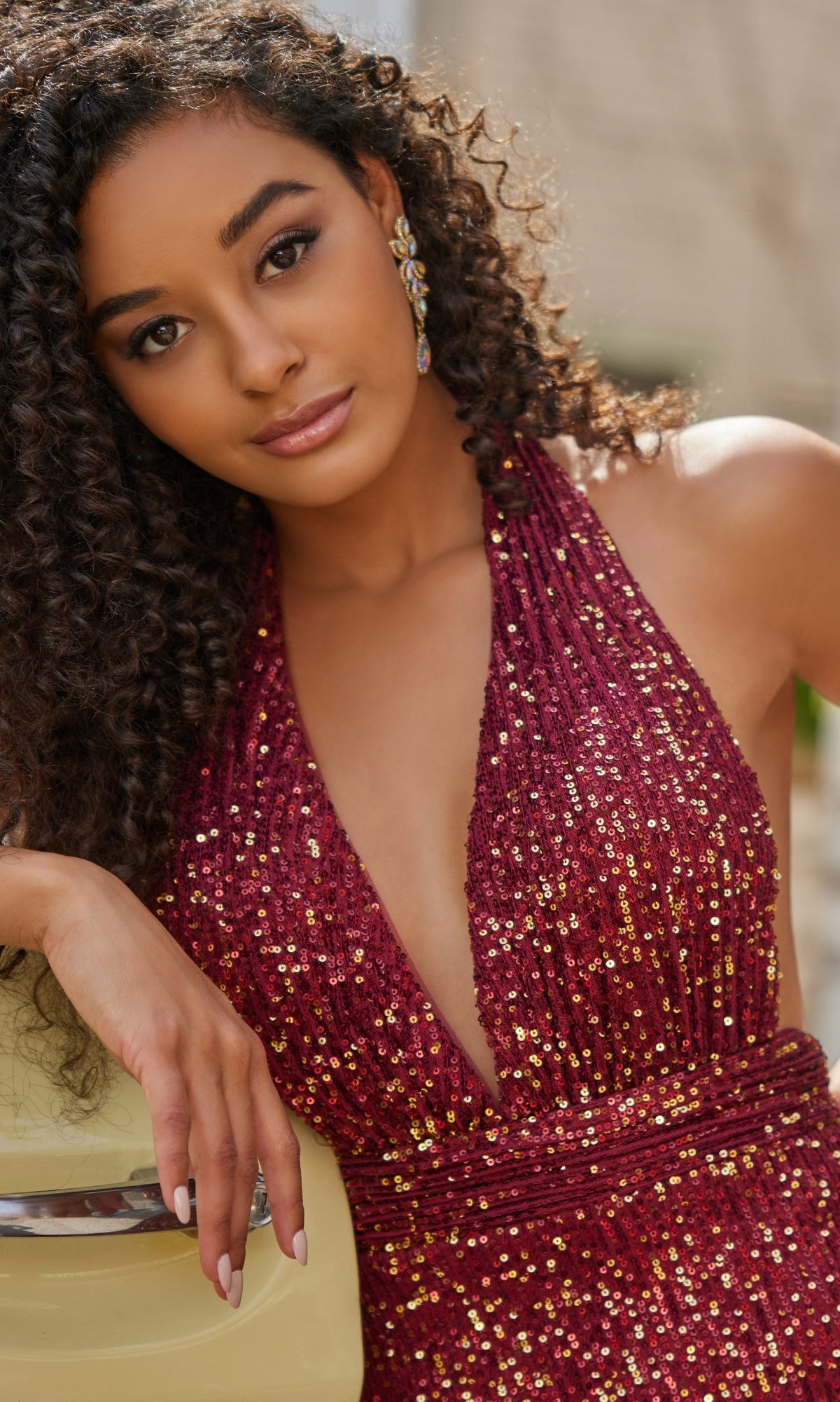 Woman with curly hair wears the Colette Long Sequin Halter Prom Dress CL5115 in deep red, adorned with shimmering sequins, as she leans against a yellow surface and looks at the camera with a neutral expression.