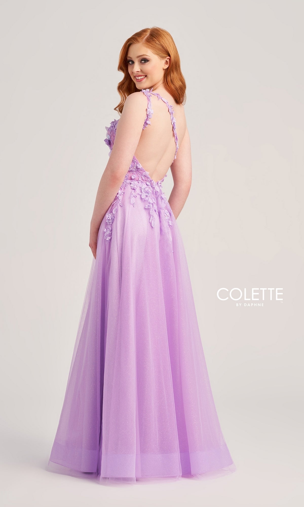 A woman with long red hair stands smiling over her shoulder, wearing the Colette One-Shoulder Designer Prom Dress CL5124—a floor-length lavender gown with floral appliqué and a backless design.