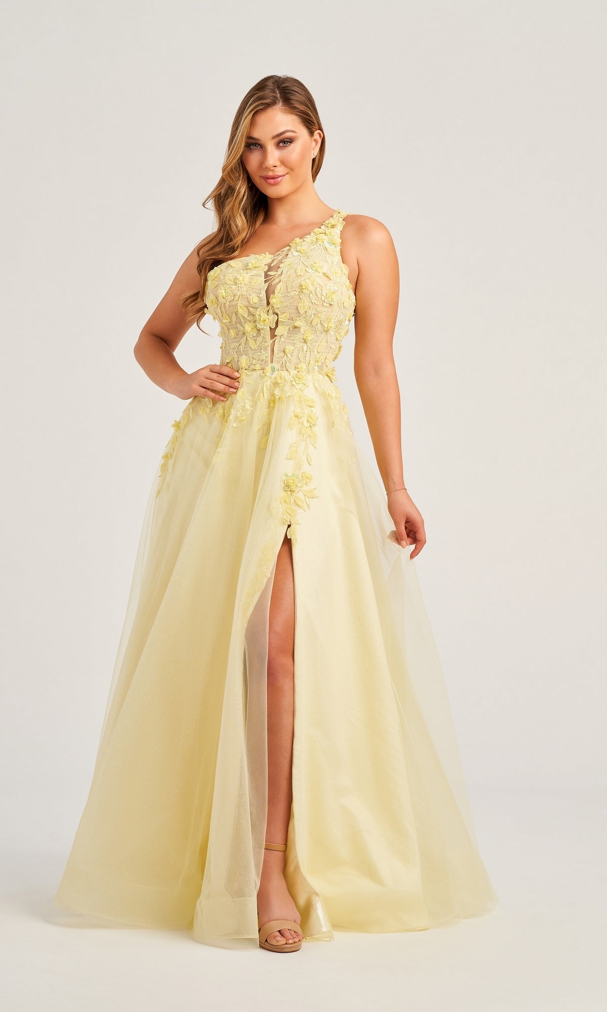 A woman poses against a plain background wearing the Colette One-Shoulder Designer Prom Dress CL5124—a floor-length yellow gown with floral appliqué details and a front slit.
