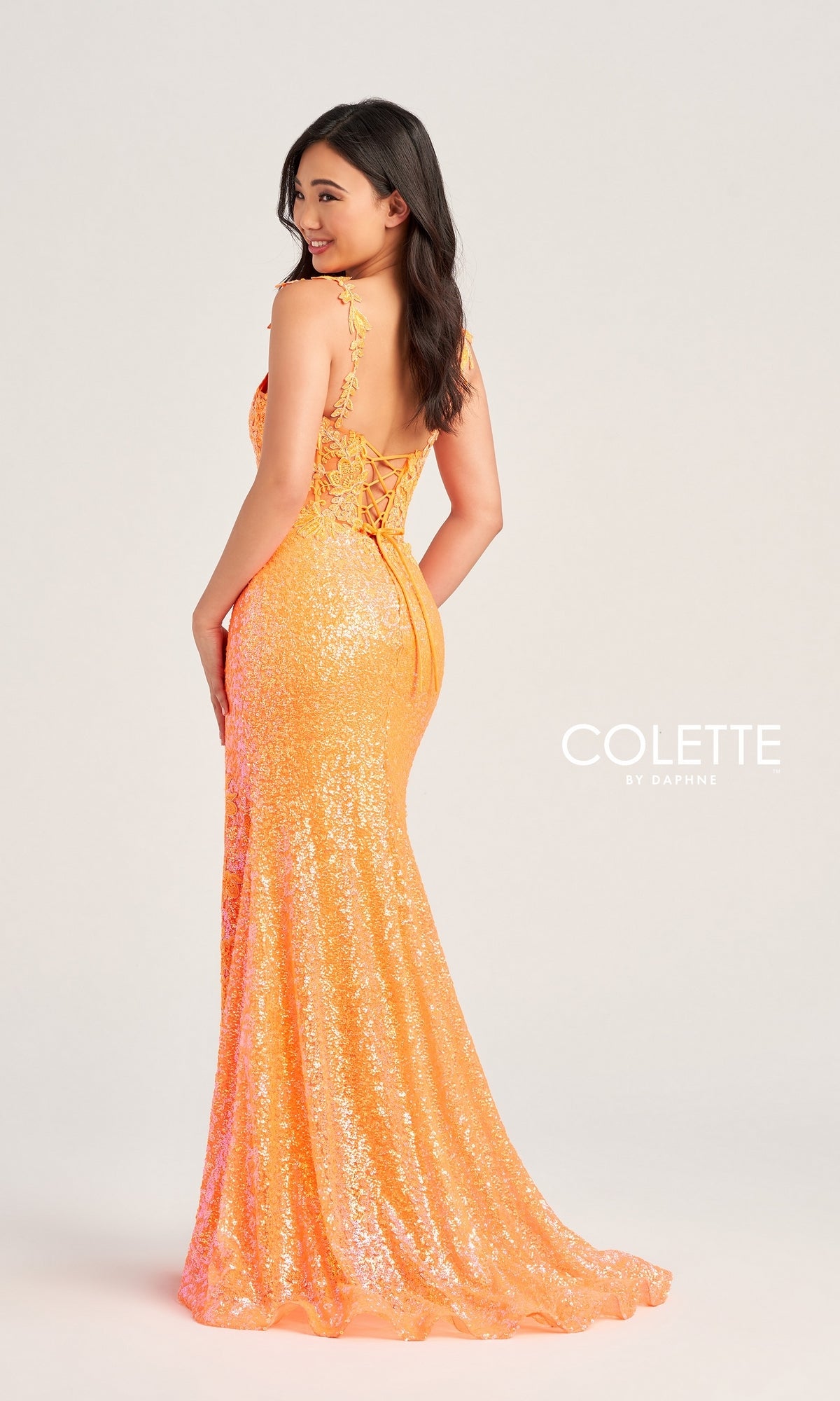 A woman poses in a floor-length, fitted orange sequin gown with a lace-up back and beaded lace appliques against a plain background. The dress is the Colette Long Sequin-Lace Prom Dress CL5133.
