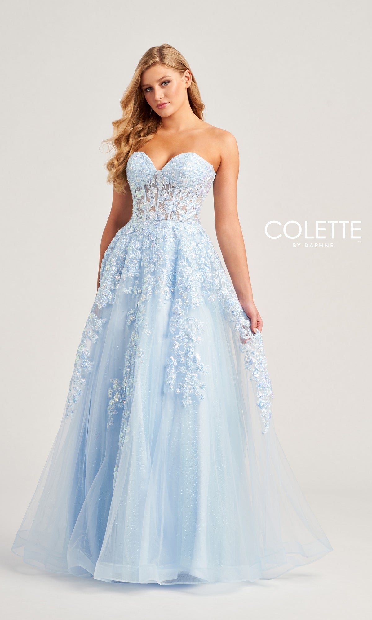 A woman in the Colette Strapless Sweetheart Prom Ball Gown CL5136—a light blue, floor-length dress with sparkly lace applique and a tulle skirt—stands against a plain background.