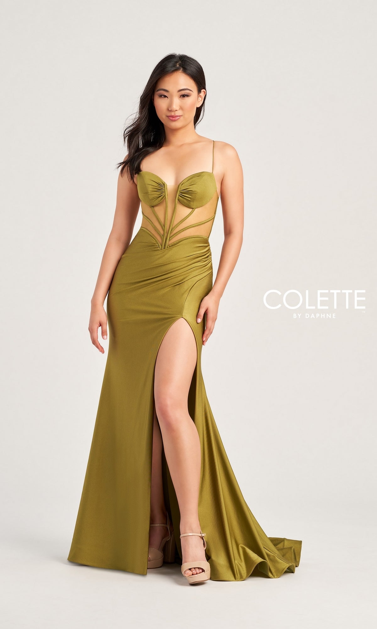 A woman in the Backless Colette Long Designer Prom Dress CL5140, olive green with a sheer bodice, spaghetti straps, and a high slit, pairs it with nude platform heels against a plain background.