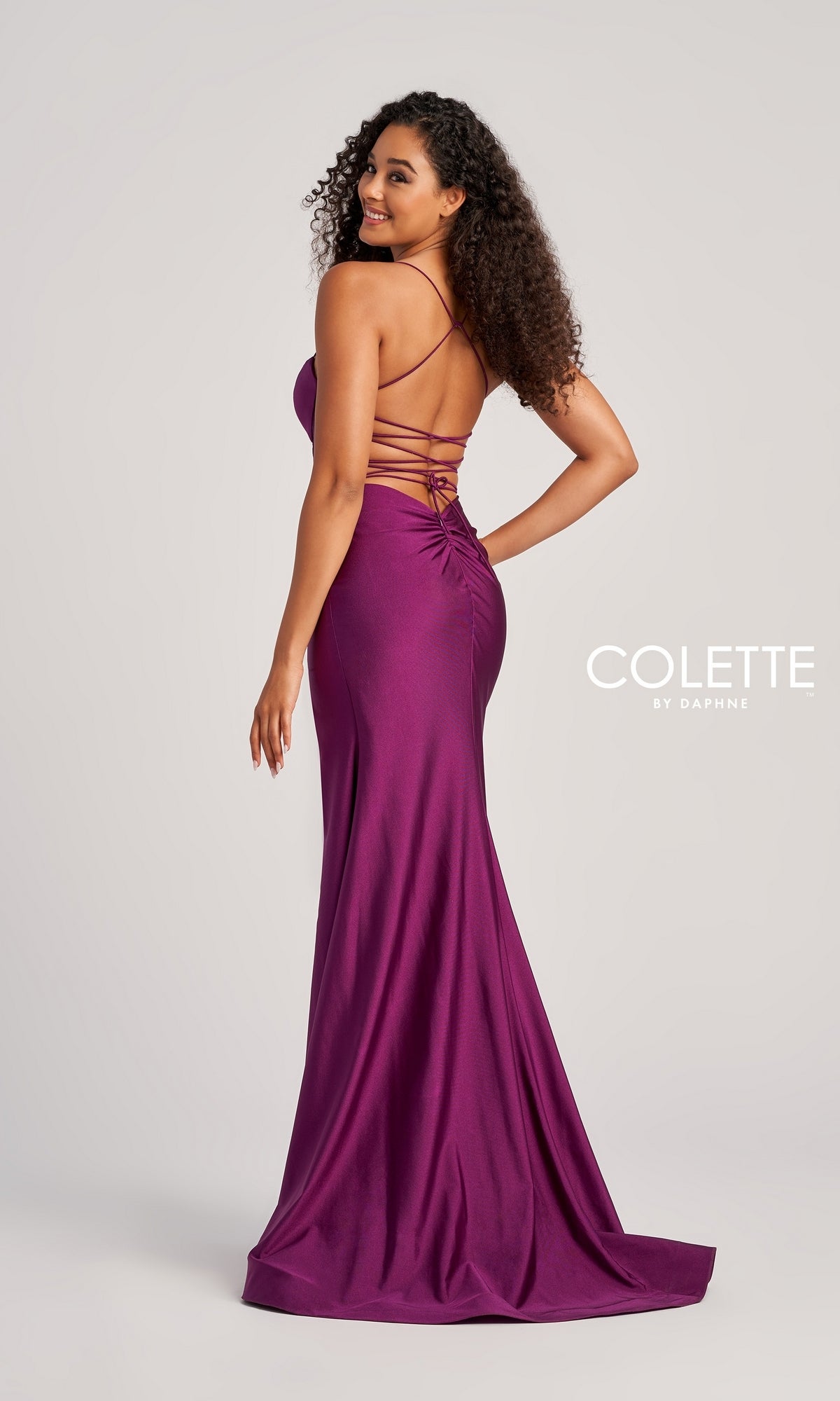 A woman stands smiling over her shoulder in the Backless Colette Long Designer Prom Dress CL5140—a floor-length, sleeveless magenta gown with a lace-up open back—against a plain background, with “COLETTE by Daphne” visible.