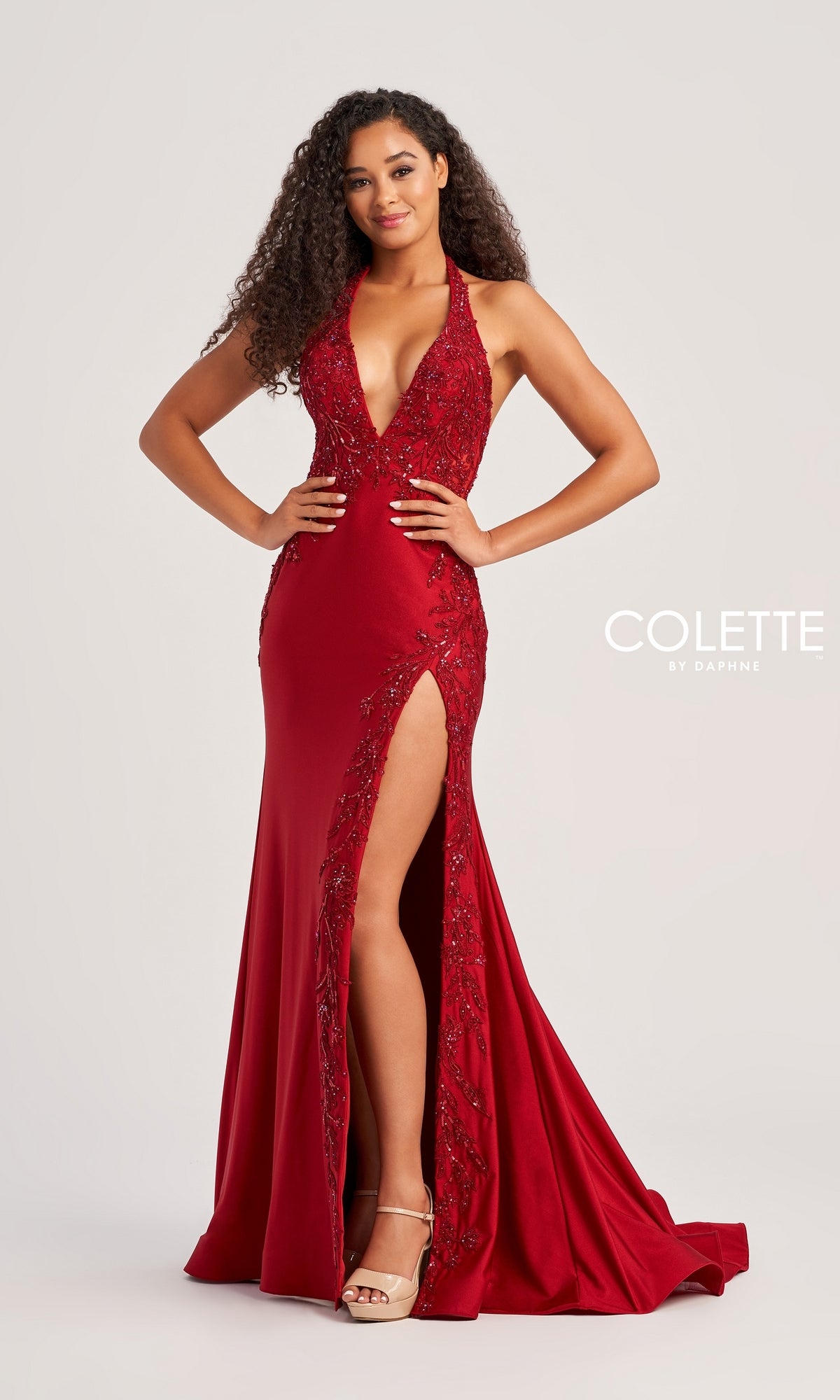 A woman poses confidently in the Lace-Bodice Colette Long Halter Prom Dress CL5206, a floor-length deep red gown with sparkly lace detailing and a high slit, styled with nude heels.