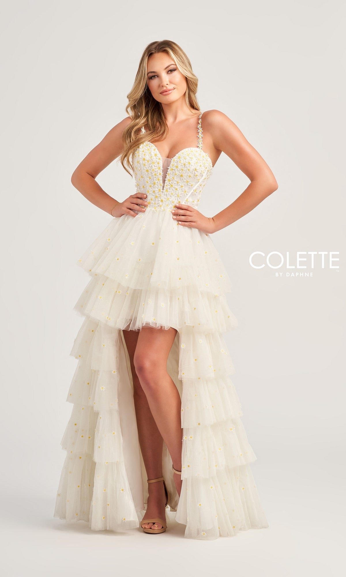 A woman poses in the Colette High-Low Daisy Tiered Prom Dress CL5237, a white prom dress with yellow embellishments and spaghetti straps, against a plain light background.