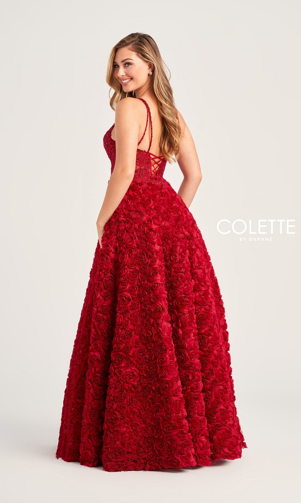 A woman smiles as she stands sideways in the Colette Long Red Rosette Prom Dress CL5251, a sleeveless, floor-length gown with 3-D rosettes and a lace-up back.