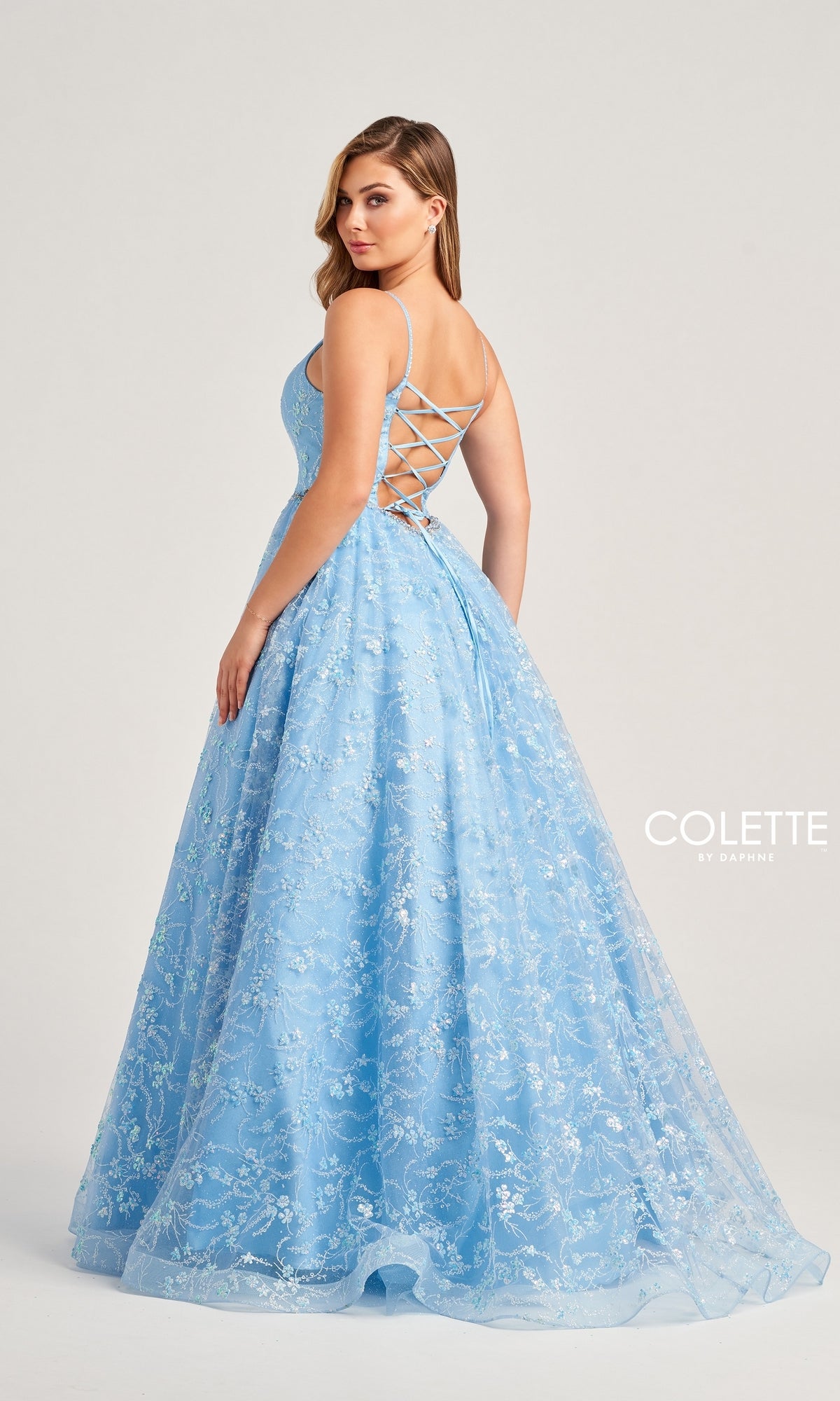 A woman stands facing away, looking over her shoulder in the Glitter-Tulle Colette Long Prom Ball Gown CL5288—a light blue, floor-length dress with floral patterns, a lace-up back, a shimmering full skirt, and glittery tulle fabric.