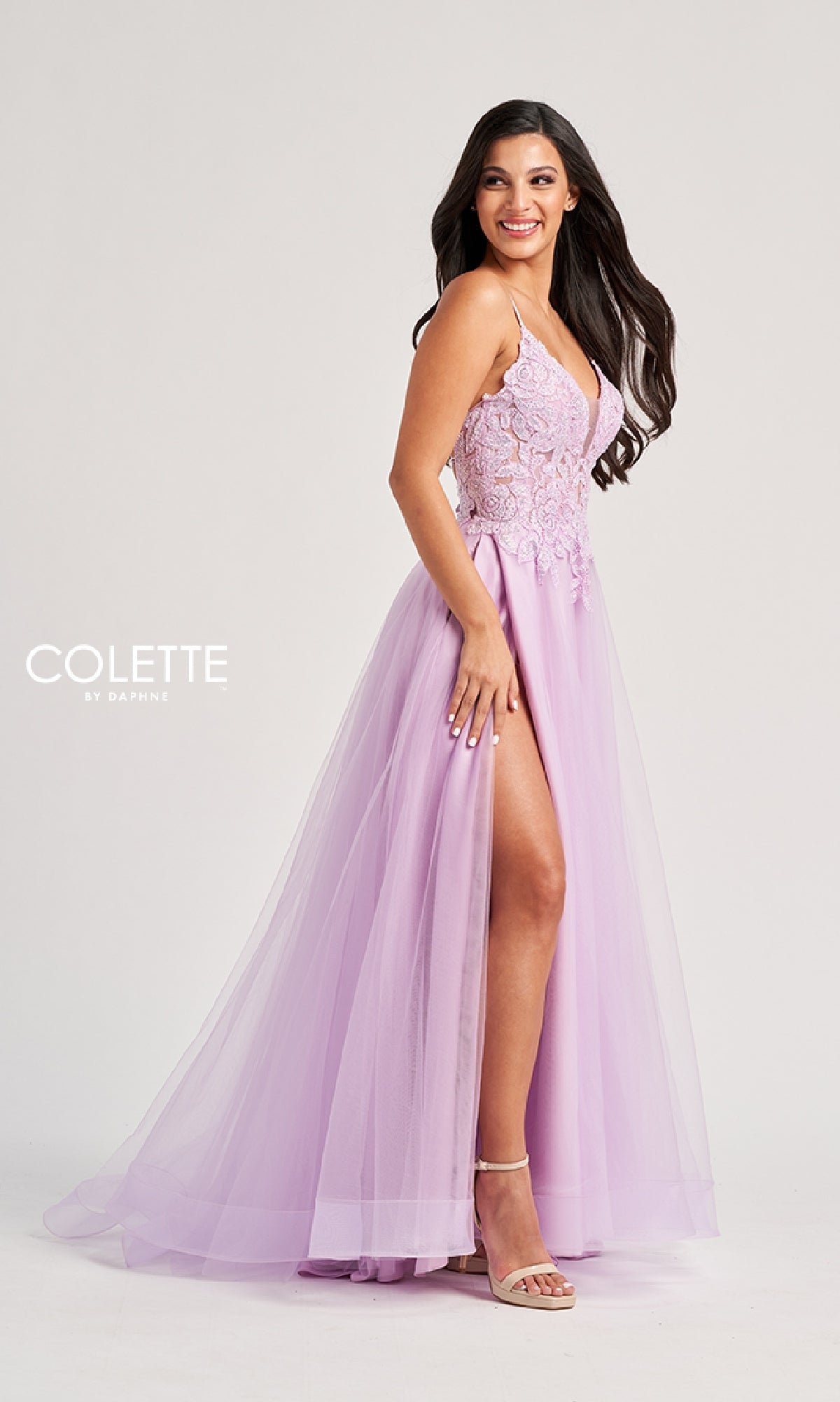 Woman in the Colette CL8100 long prom dress, featuring a sleeveless design, lace-embroidered bodice, sheer tulle skirt with a high slit, standing sideways and smiling with one leg showing.