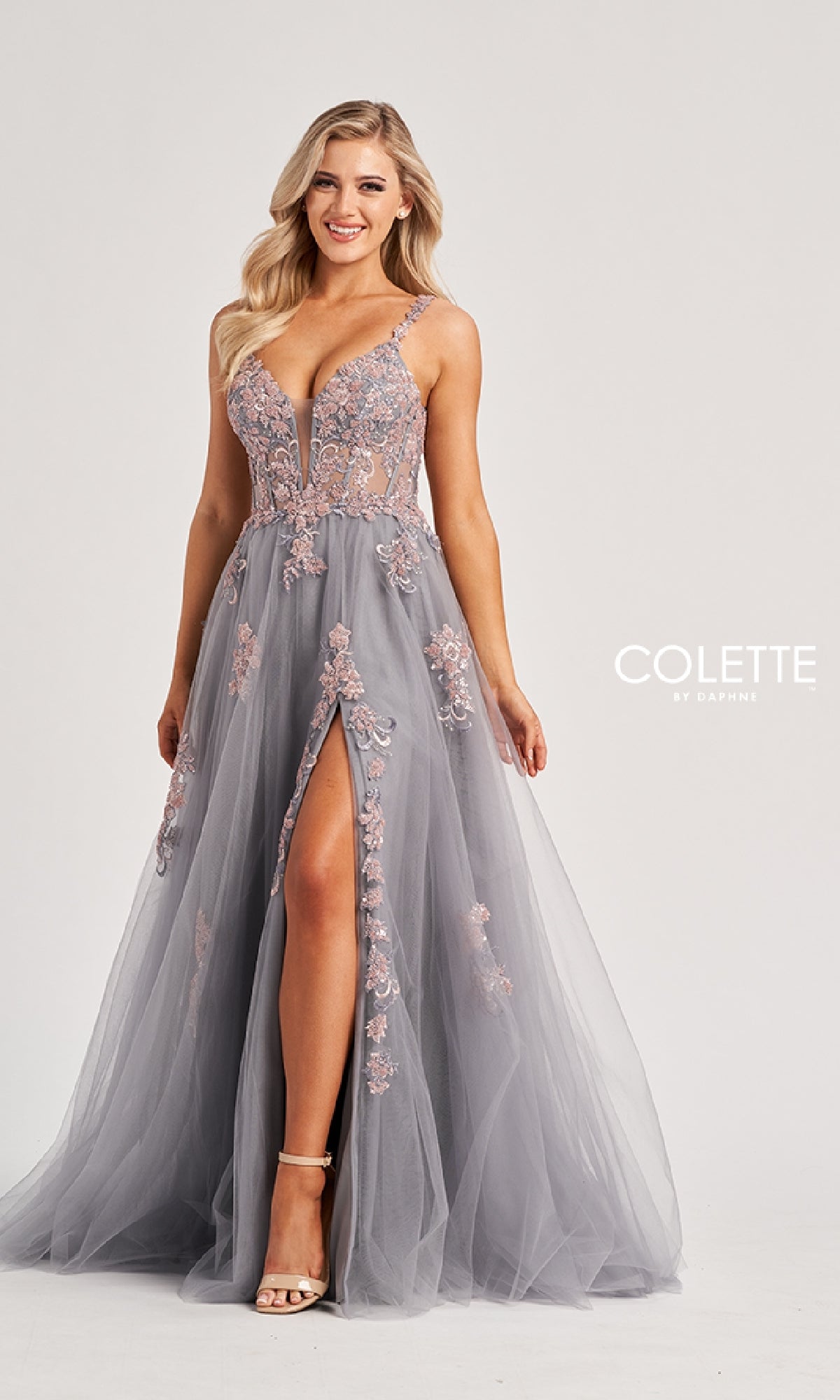 A woman models the Colette CL8130 long prom dress, featuring gray fabric with pink floral embroidery, a sheer bodice, deep V-neckline, high leg slit, and lace-up strappy open back against a plain white background.