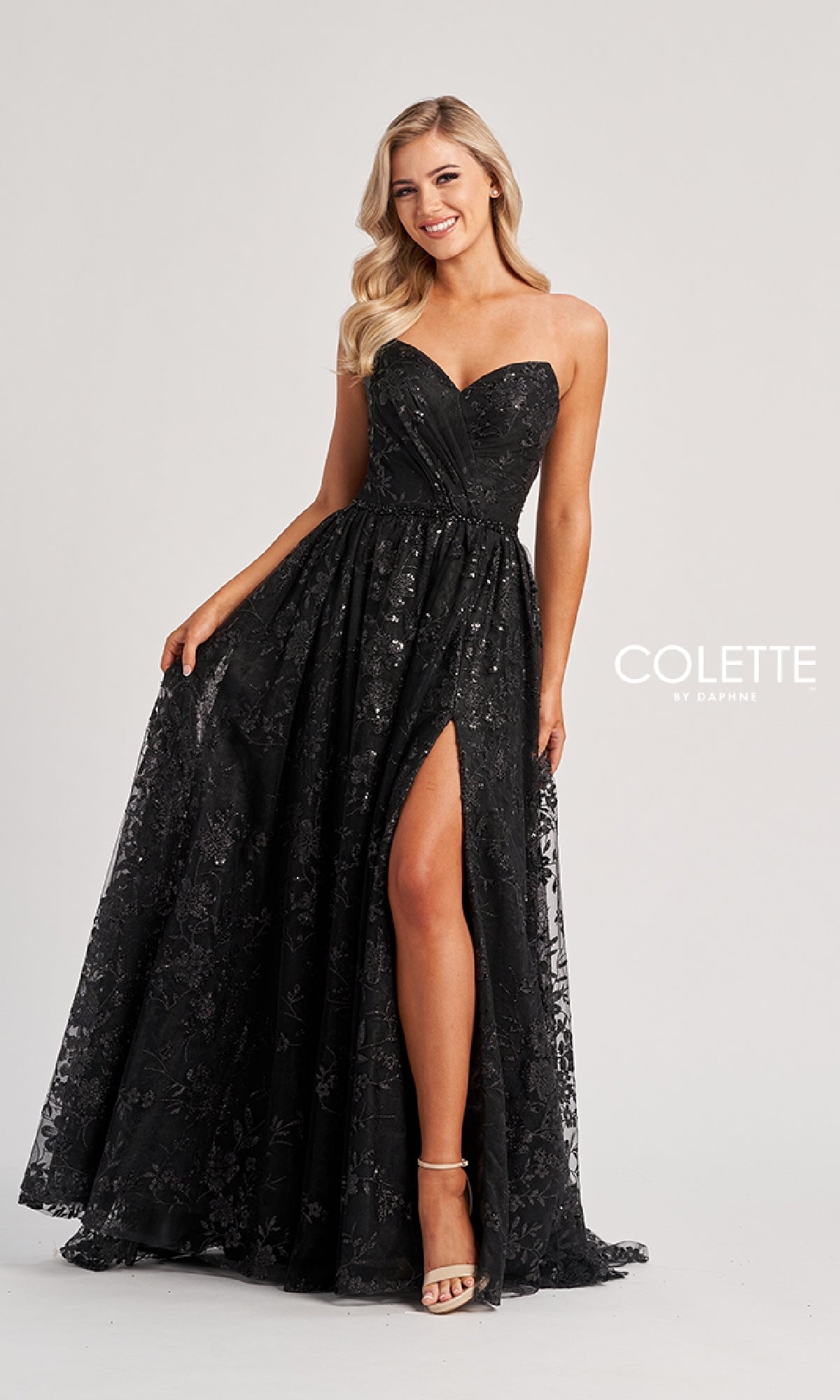 A woman wearing the Colette CL8260 long black strapless prom dress with a thigh-high slit and floral sequins smiles against a plain background.