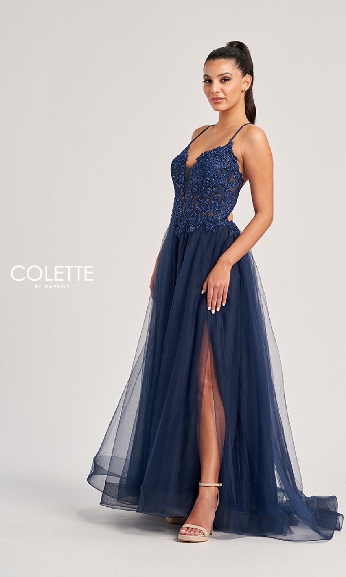 A woman poses against a plain background wearing the Colette CL8320, a navy blue long prom dress with an embroidered bodice, spaghetti straps, and an A-line tulle skirt featuring a high slit.