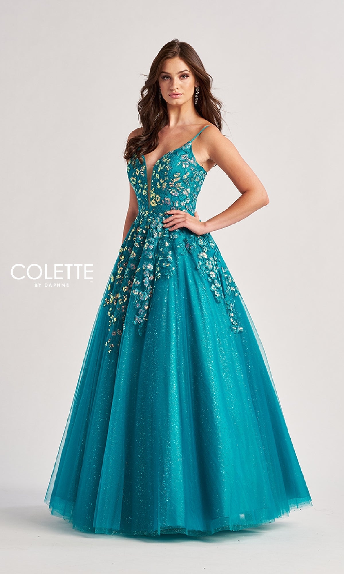 A woman in the Colette CL8420 Low V-Neck Glitter Prom Ball Gown, featuring a teal floor-length sleeveless design with floral embroidery and delicate embellishments, stands with one hand on her hip against a plain background.